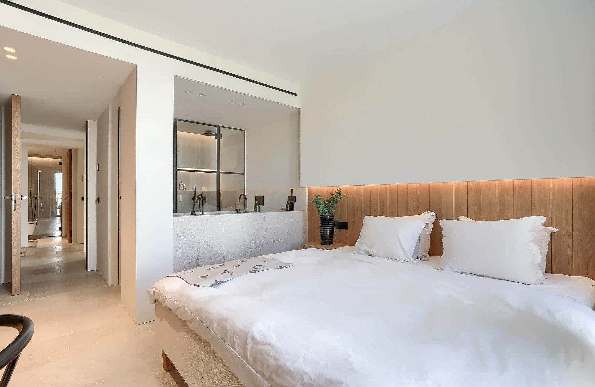 Modern bedroom with a white bed, wood-paneled headboard, and an open-concept en-suite bathroom featuring marble surfaces.