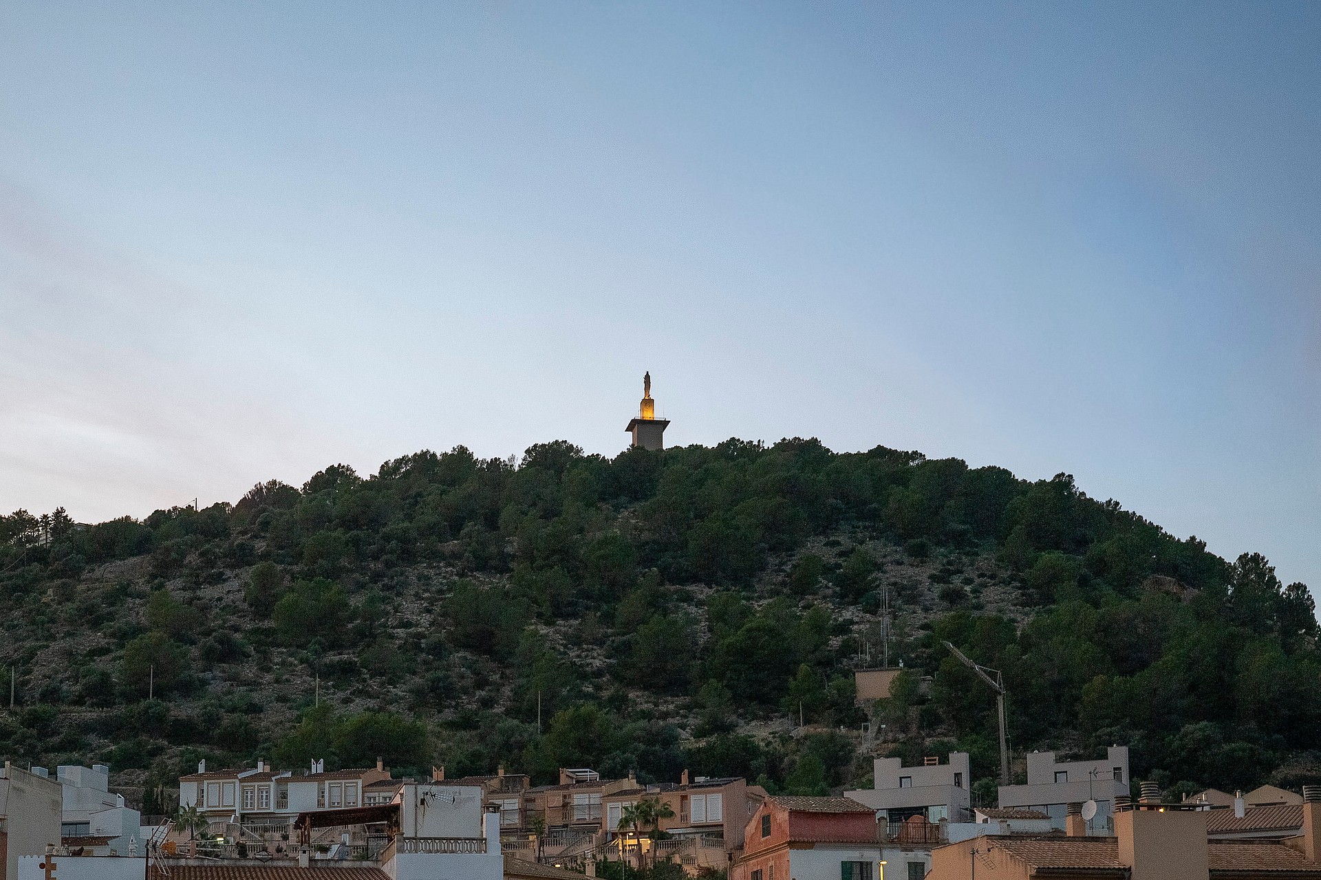 A distant view of a hillside topped with a tall landmark tower against a clear blue evening sky, seen above local rooftops.