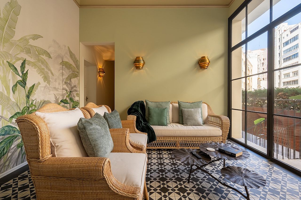 Interior sunroom with woven rattan furniture, a large palm leaf mural, and black-framed glass windows overlooking the city.