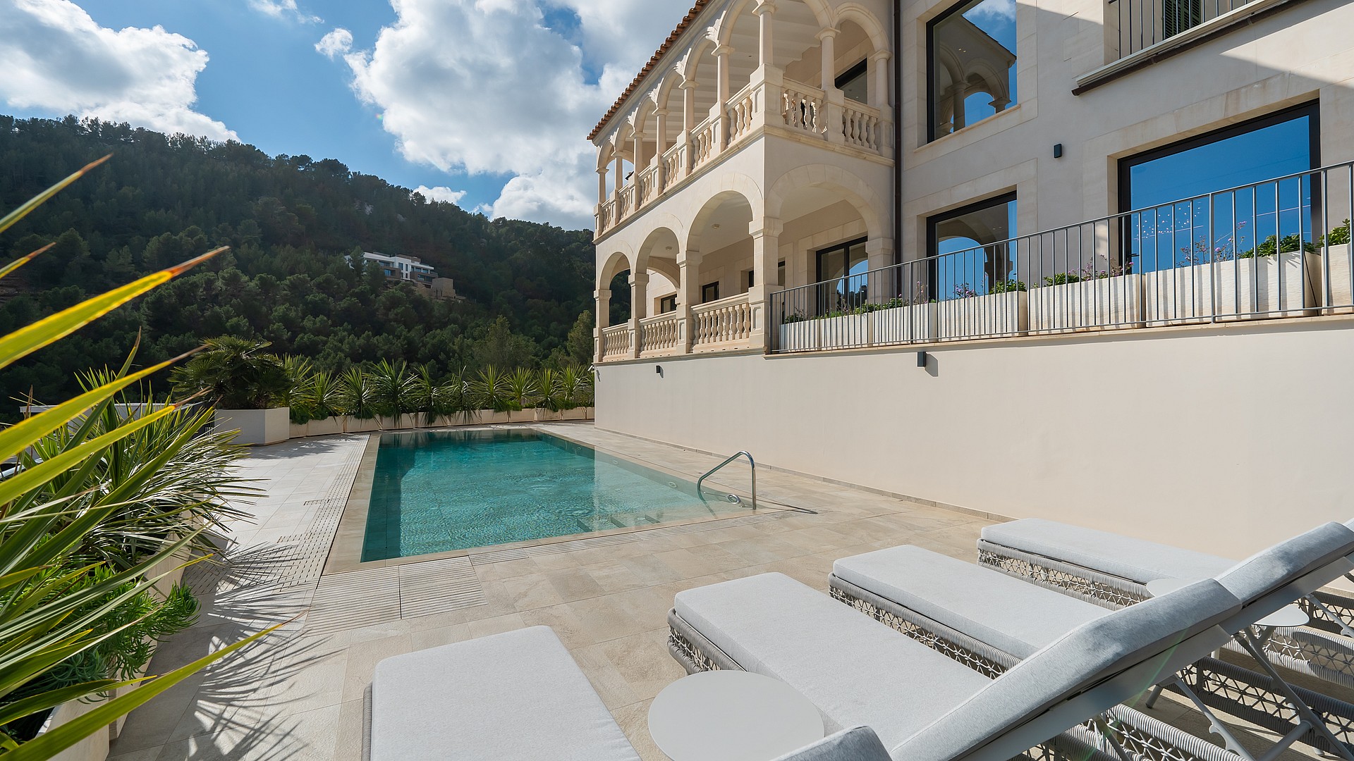 A rectangular swimming pool on a tiled terrace with white sun loungers, set against a multi-story villa with arched balconies.