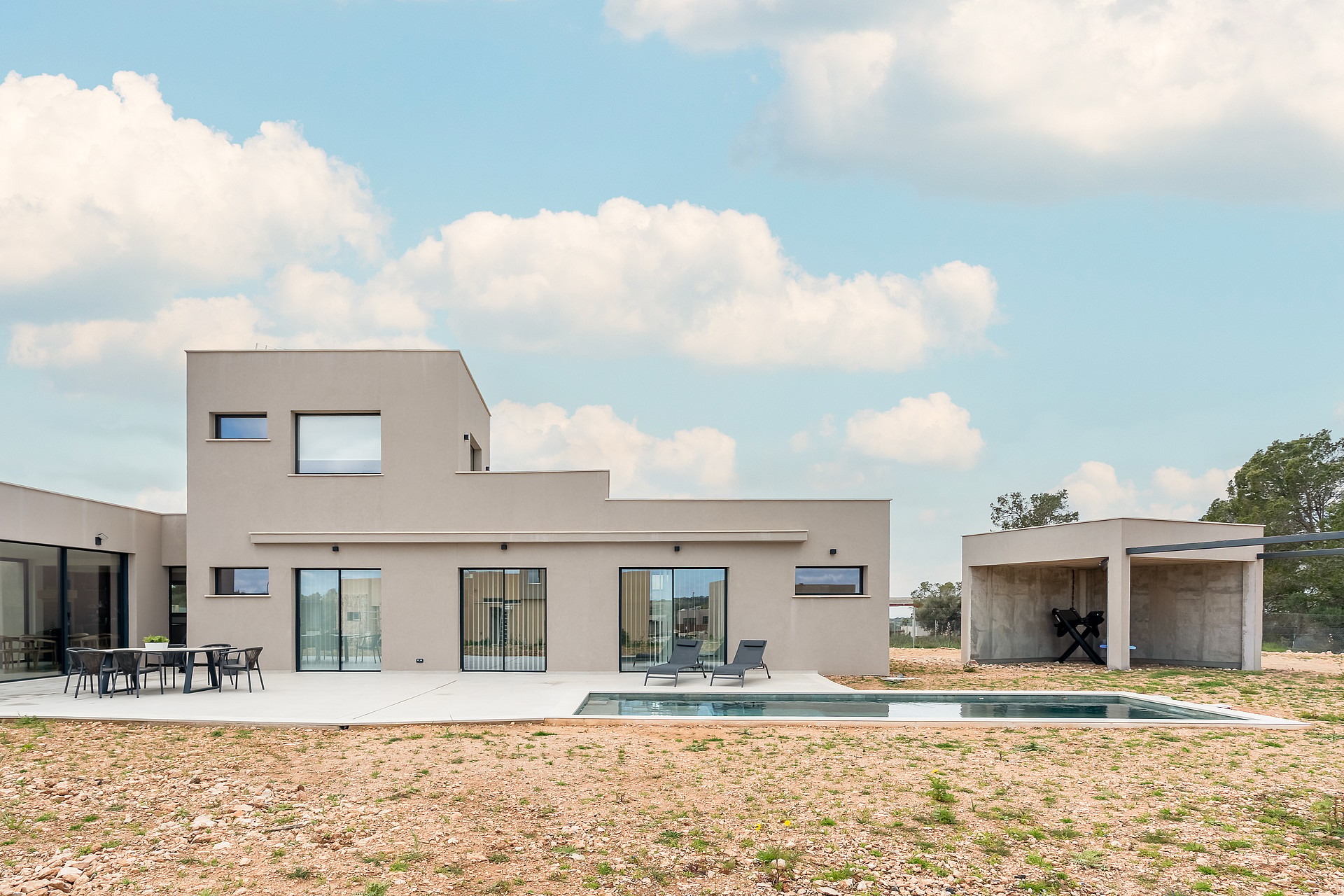 Rear view of a modern two-story beige villa with a swimming pool, paved terrace, and a separate covered outdoor lounge area.