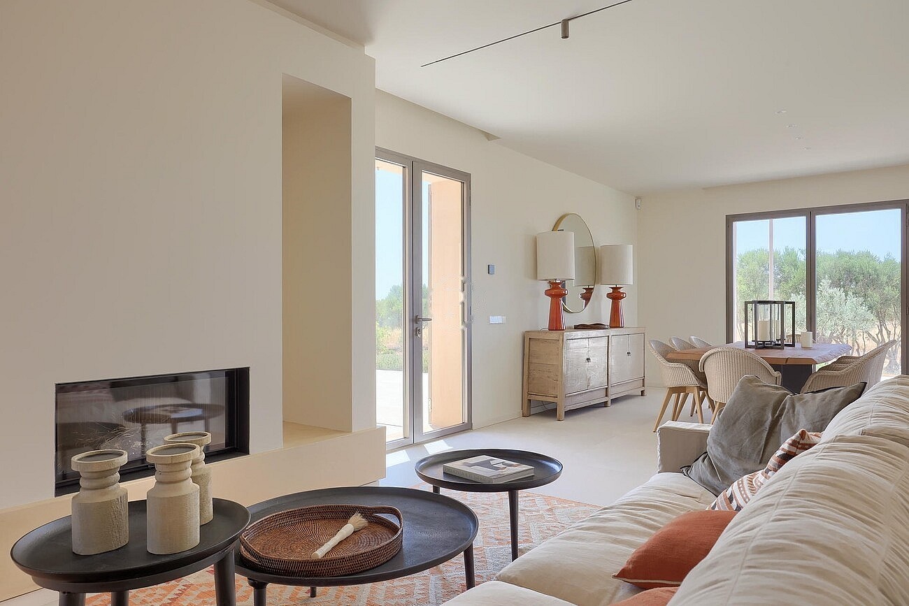 An open-plan living and dining room with a built-in fireplace, light-toned sofas, two round coffee tables, and a wooden dining table in the background, facing exterior mountain views.