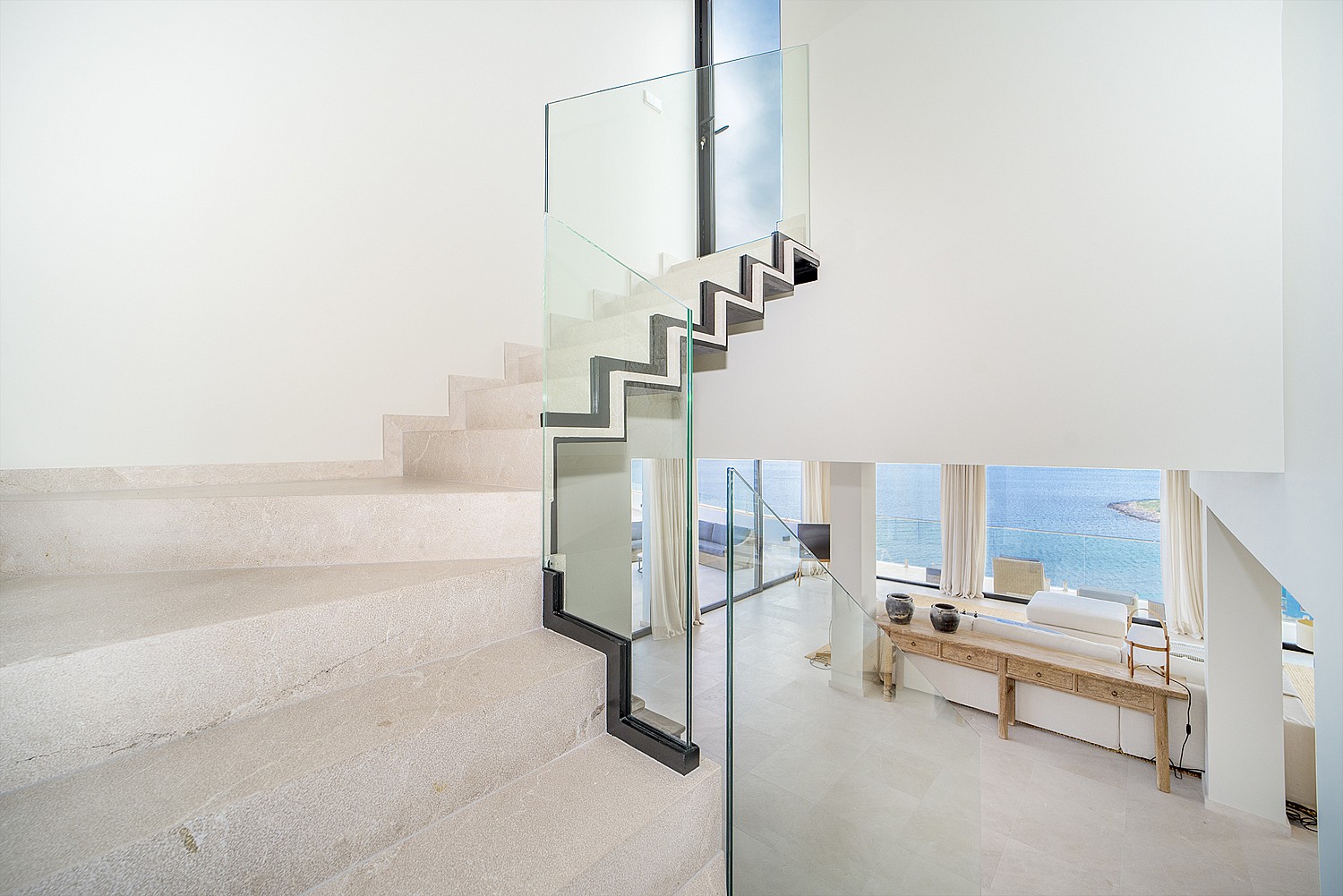 A white stone staircase with a clear glass railing connects to an upper floor, overlooking an open-plan living area with sea views.