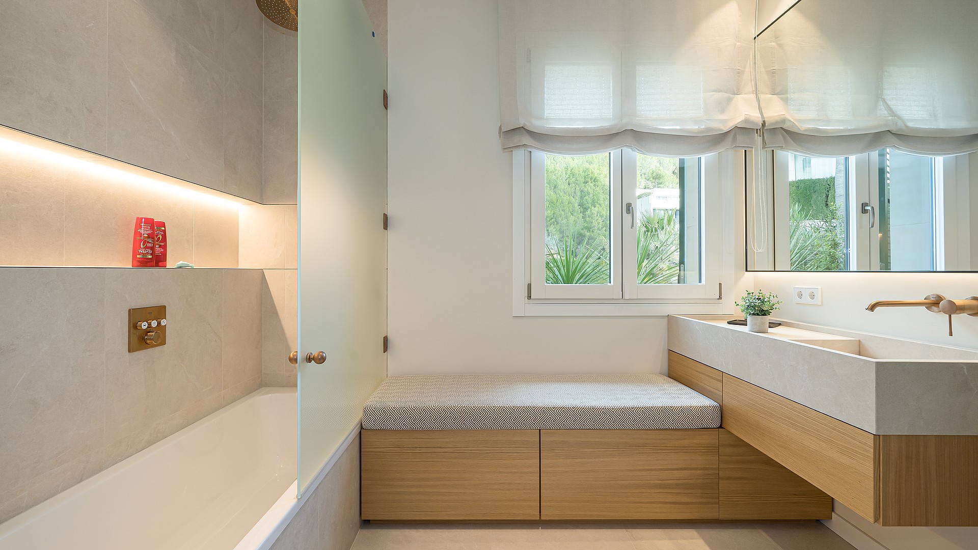Contemporary bathroom with a white built-in tub, stone-textured walls, oak wooden vanity, and a window with white Roman blinds.