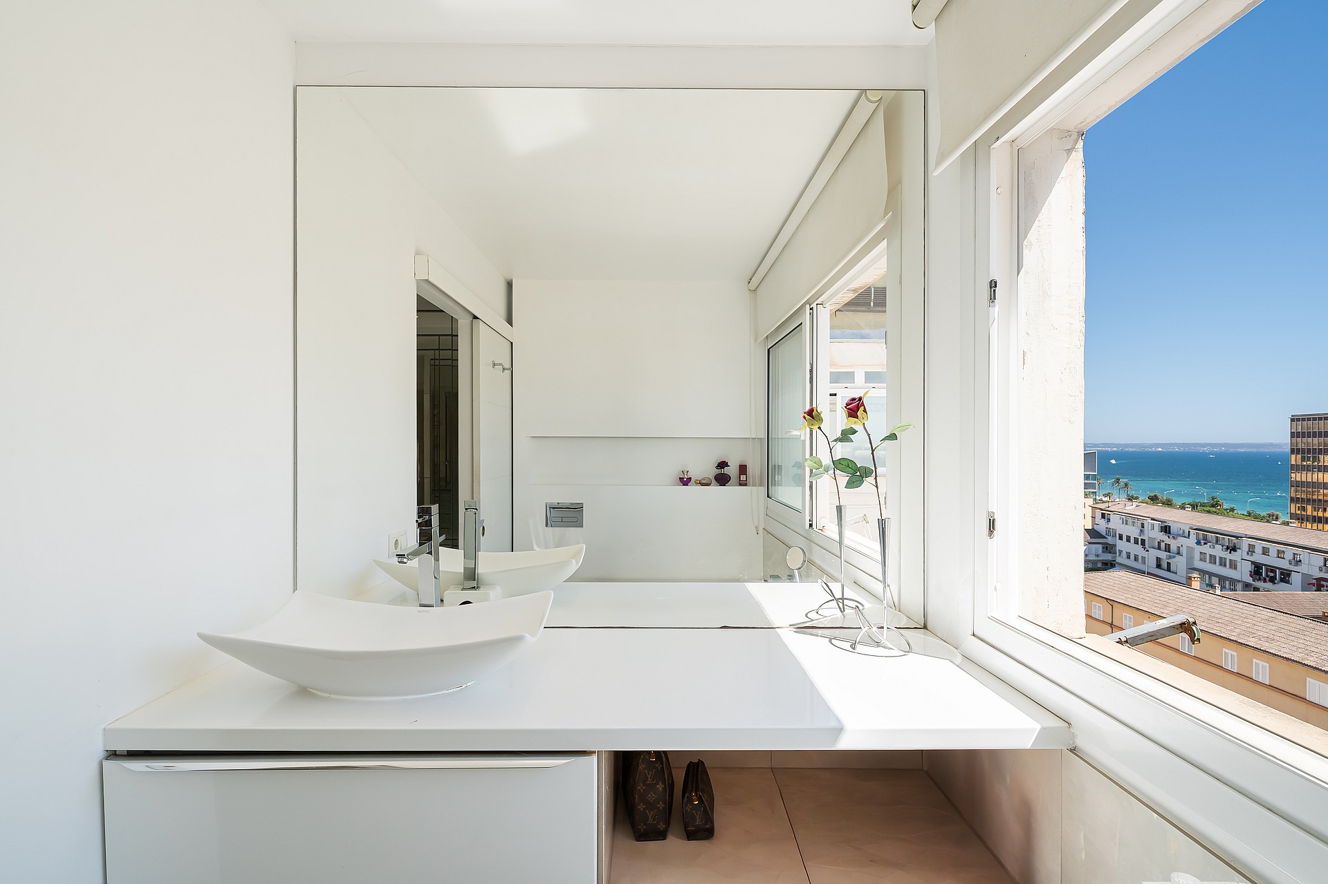 A modern white bathroom vanity with a square vessel sink and a large wall mirror overlooking a sunlit coastal city scene.
