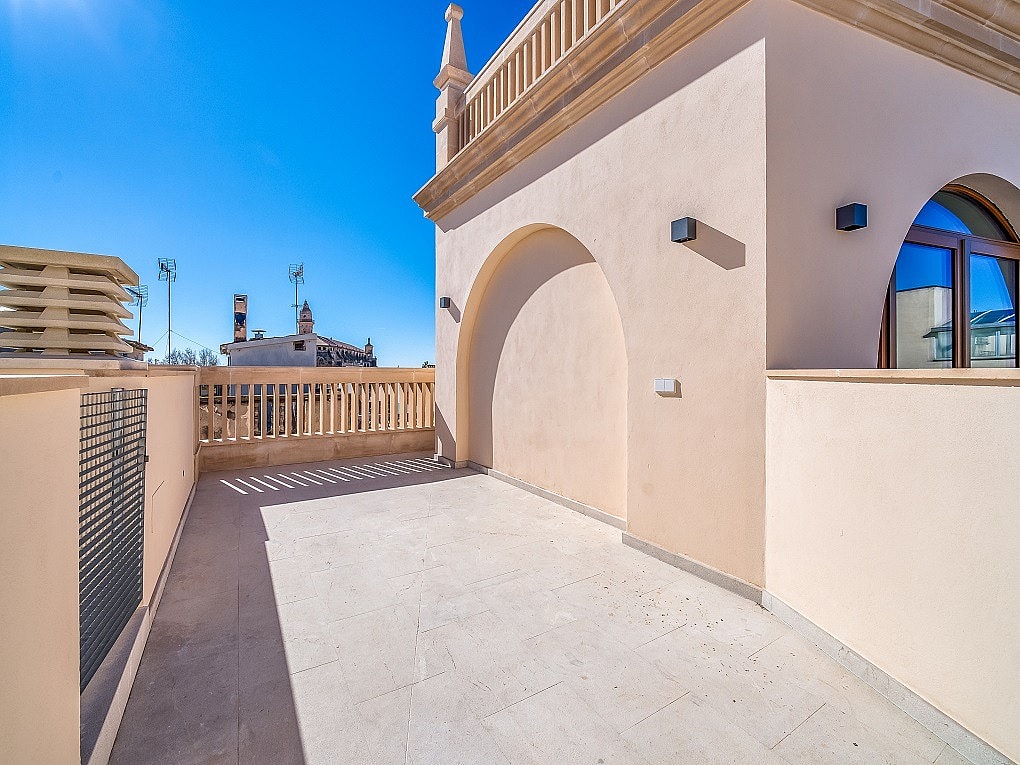 A rooftop terrace featuring light-colored stone flooring, stucco walls with a decorative arch, and stone balustrade railings under a clear blue sky.