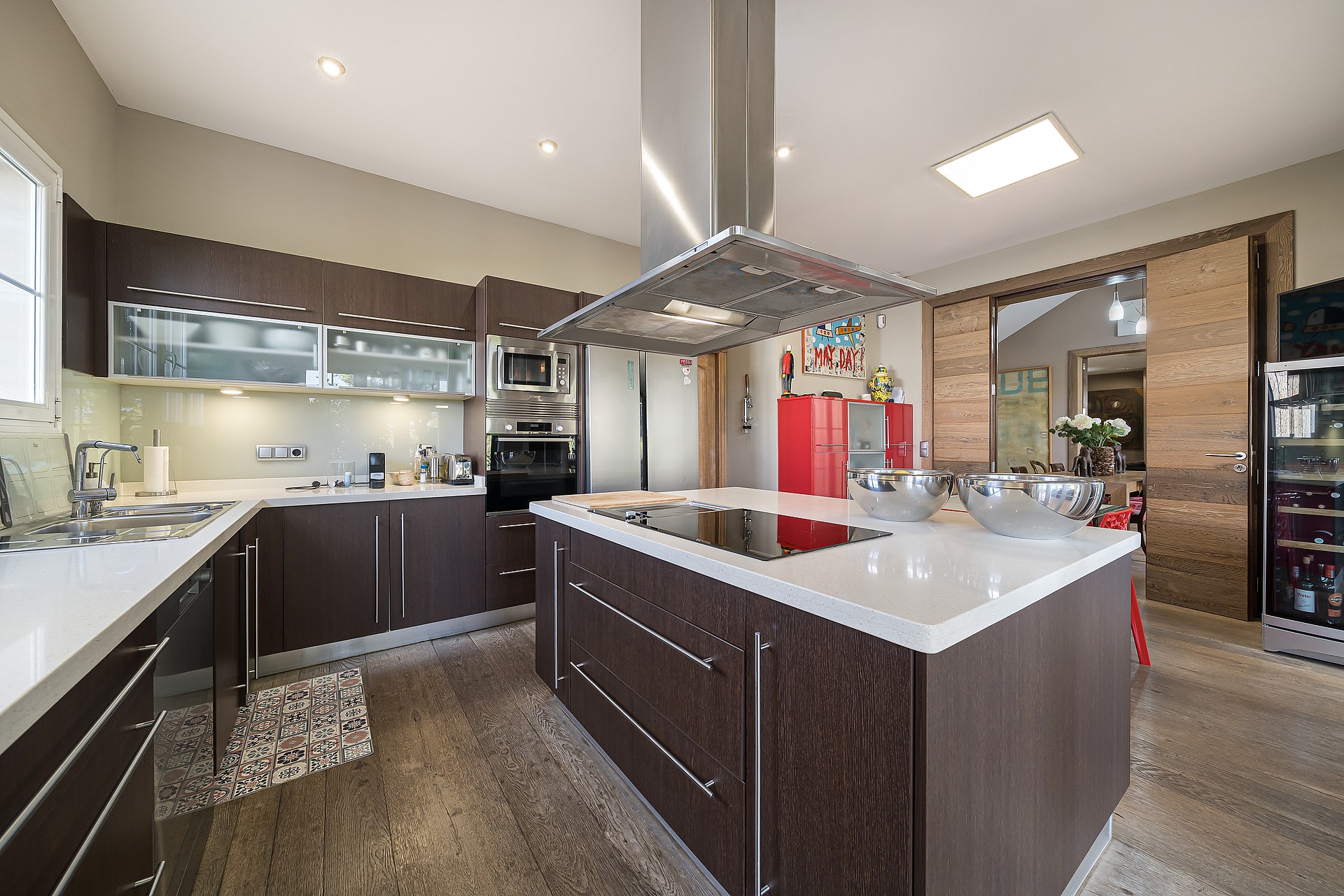 Modern kitchen featuring a large central island with white stone countertop, dark wood cabinetry, stainless steel range hood, and integrated appliances.