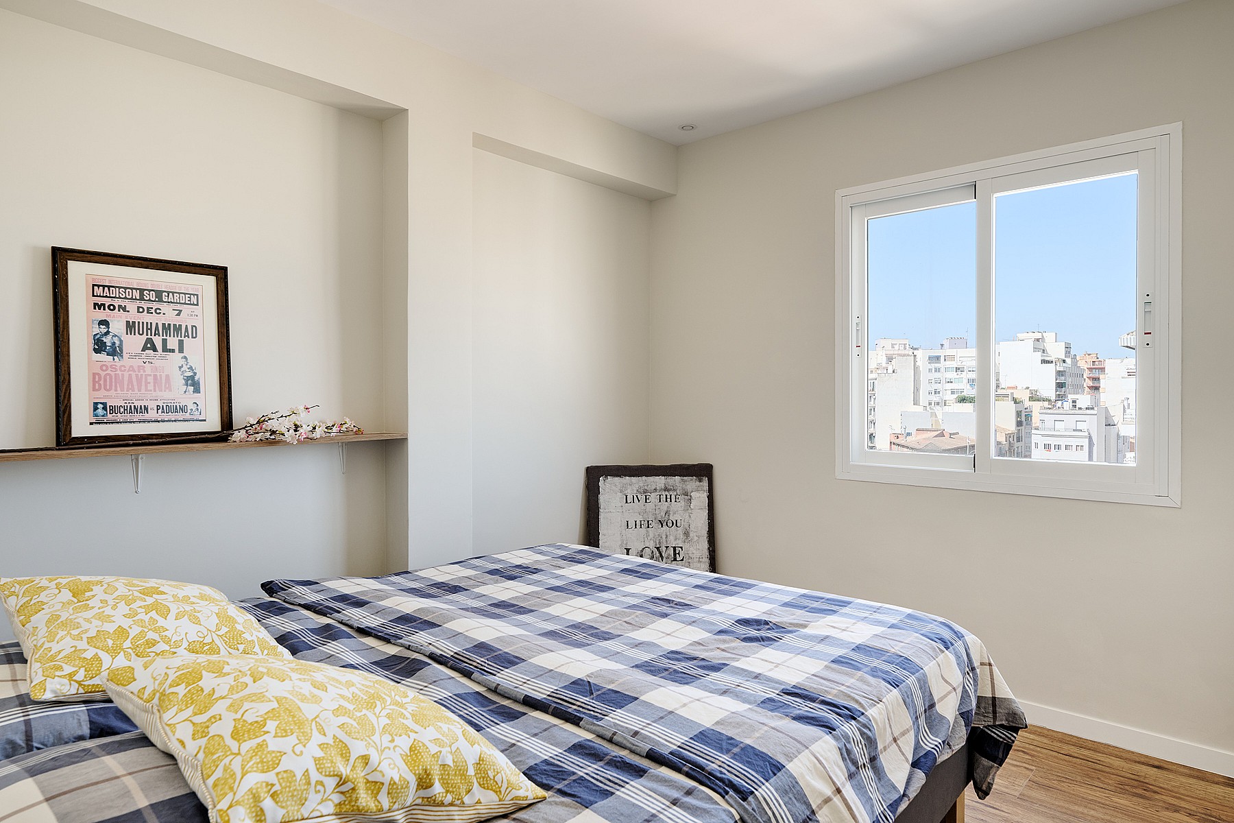 A bedroom featuring a bed with a blue plaid quilt, a floating wooden shelf with framed artwork, and a window overlooking city buildings.