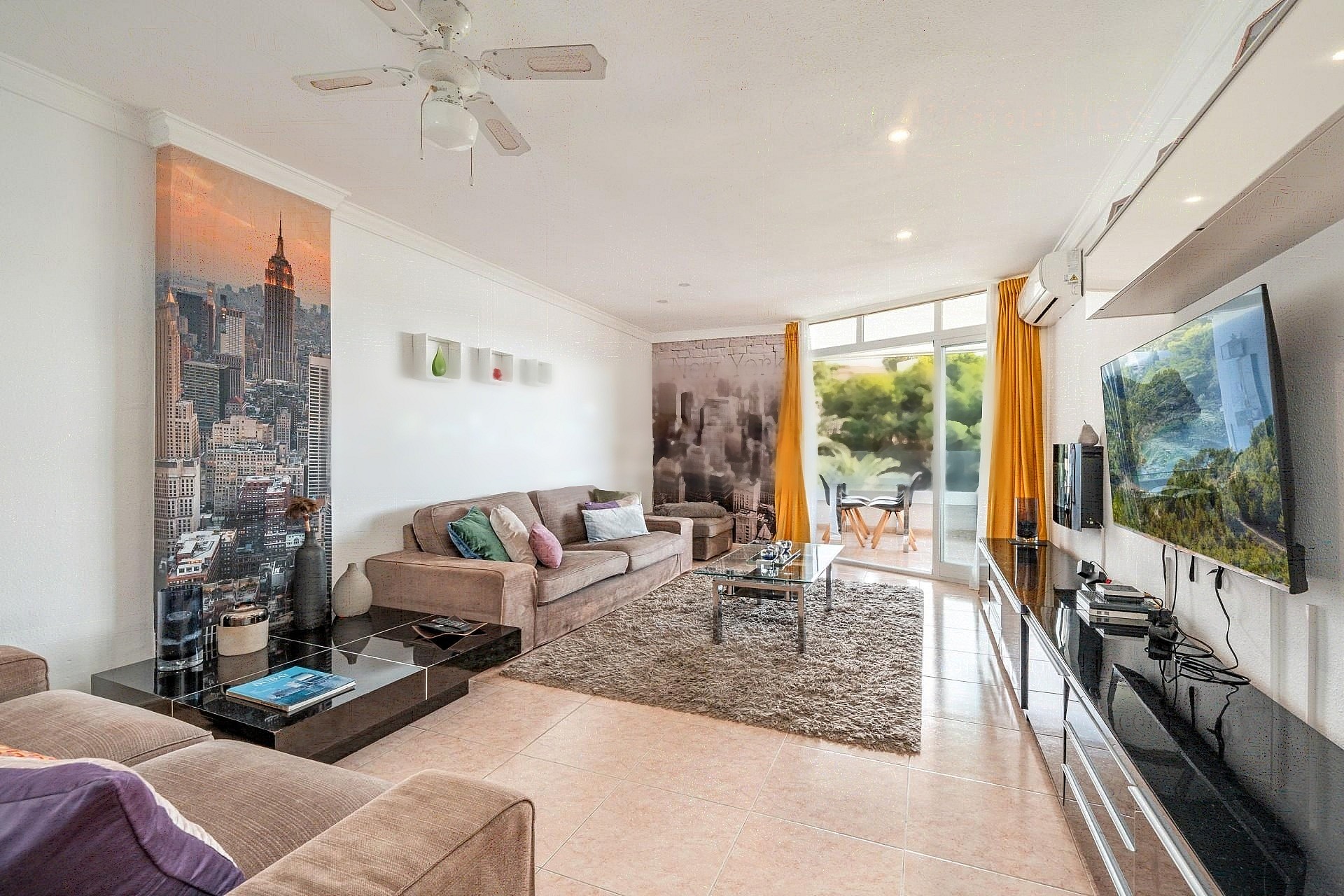 Wide-angle view of a living room featuring a plush sofa, high-gloss TV unit, and floor-to-ceiling glass doors opening to a terrace.