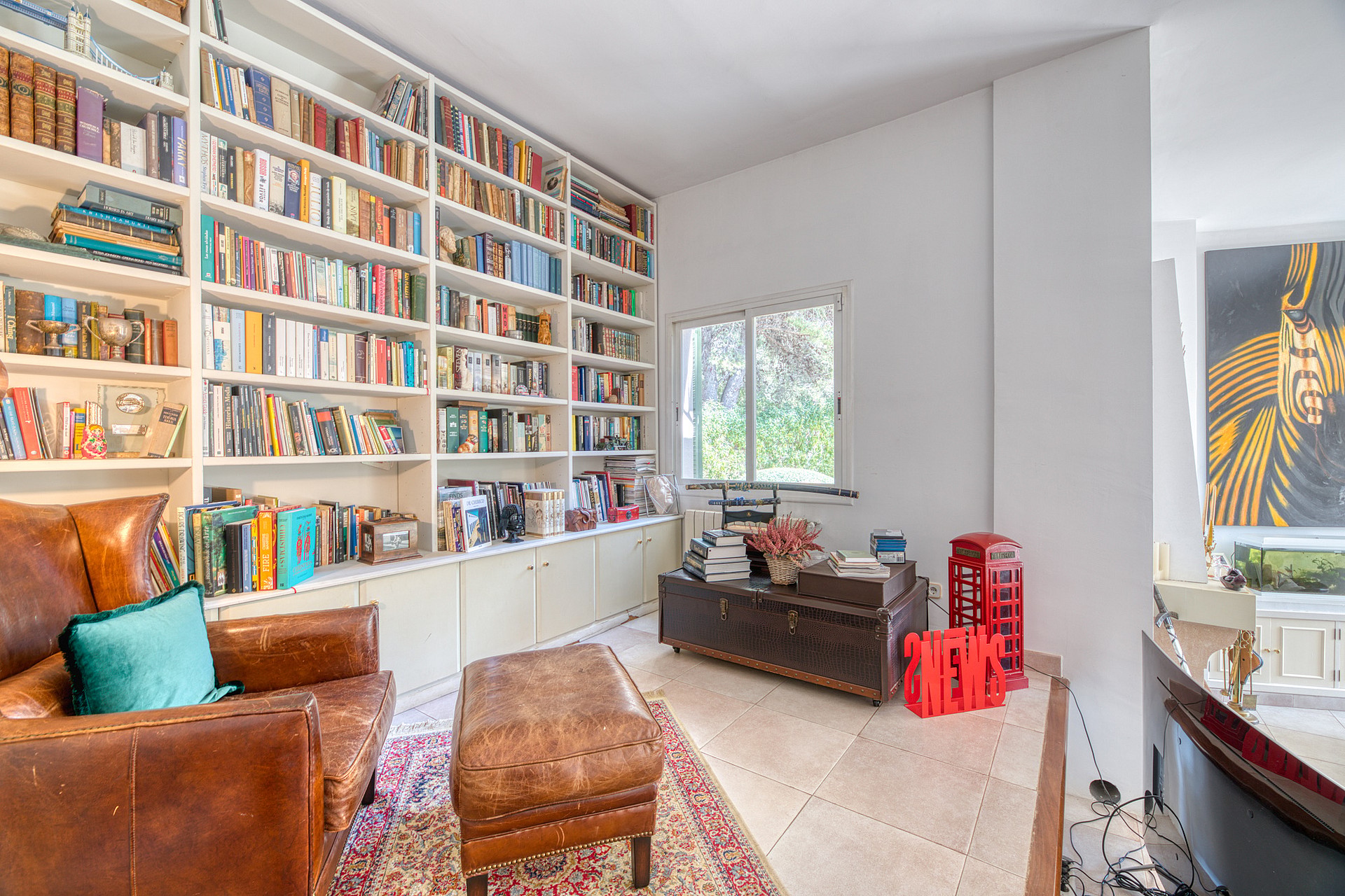 A library area with tall white bookshelves, a tan leather armchair and ottoman, a patterned rug, and a dark wooden trunk.
