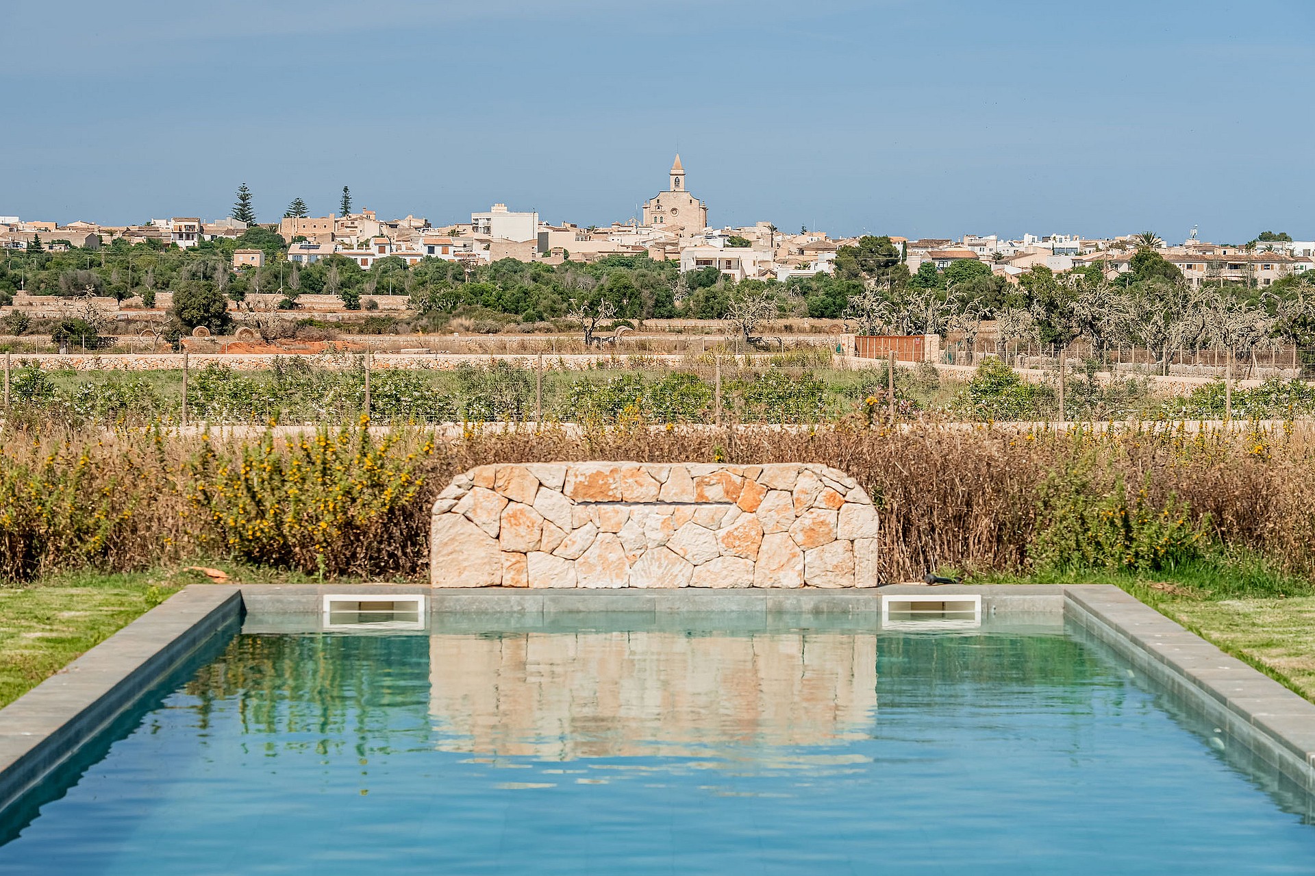 A rectangular swimming pool featuring a natural stone waterfall wall with the village of Santanyí in the background.