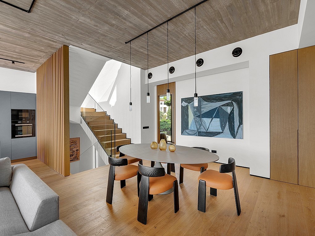 Dining area with oval table and wooden chairs, adjacent to a wooden staircase under a raw concrete ceiling.