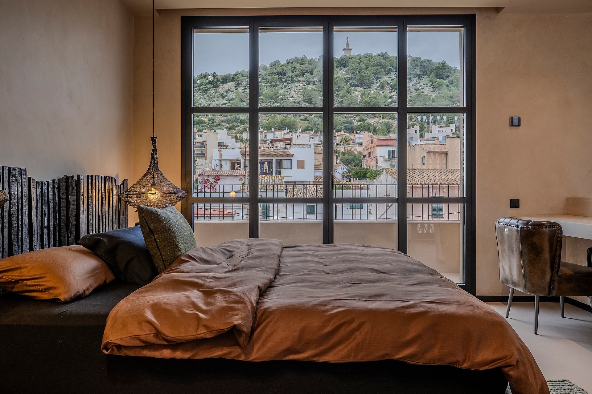 A contemporary bedroom featuring a dark bed frame with a textured wooden headboard, terracotta bedding, and a large metal-framed window looking out onto a hillside.