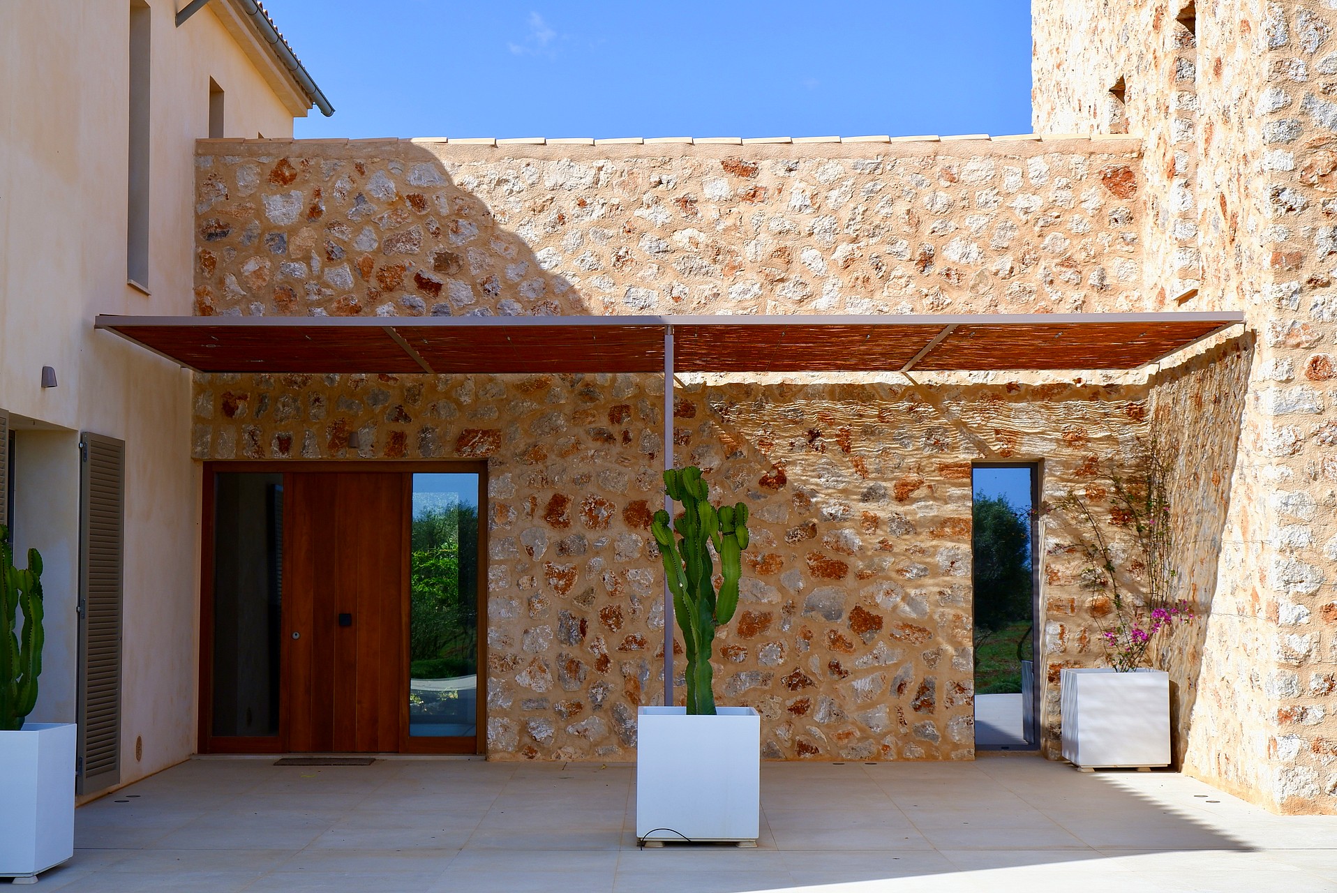 Entrance to a traditional stone-walled finca in Santanyí featuring a wooden pergola, cactus planters, and a timber door.