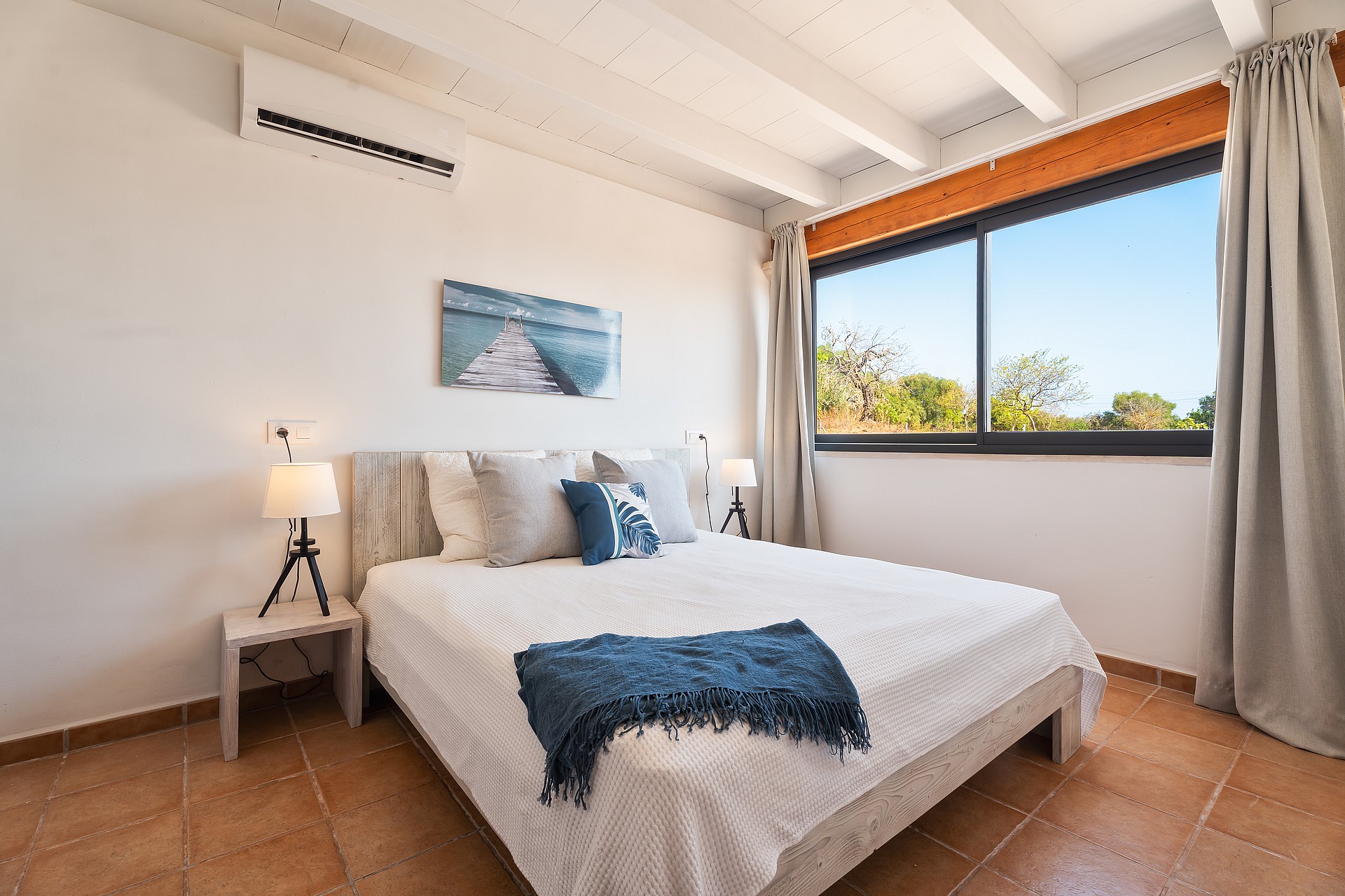 Dormitorio con paredes blancas, vigas de madera vistas, suelos de baldosas y cama frente a una ventana.