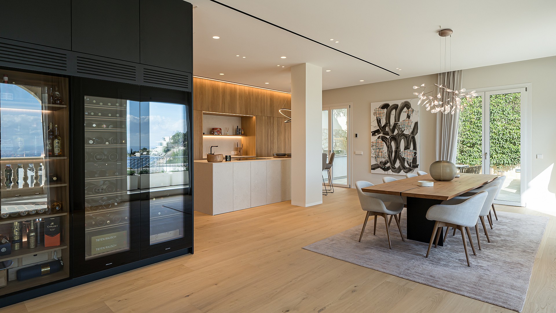 Open-plan dining area with a long wooden table, modern chandelier, and floor-to-ceiling glass wine fridge in a villa.