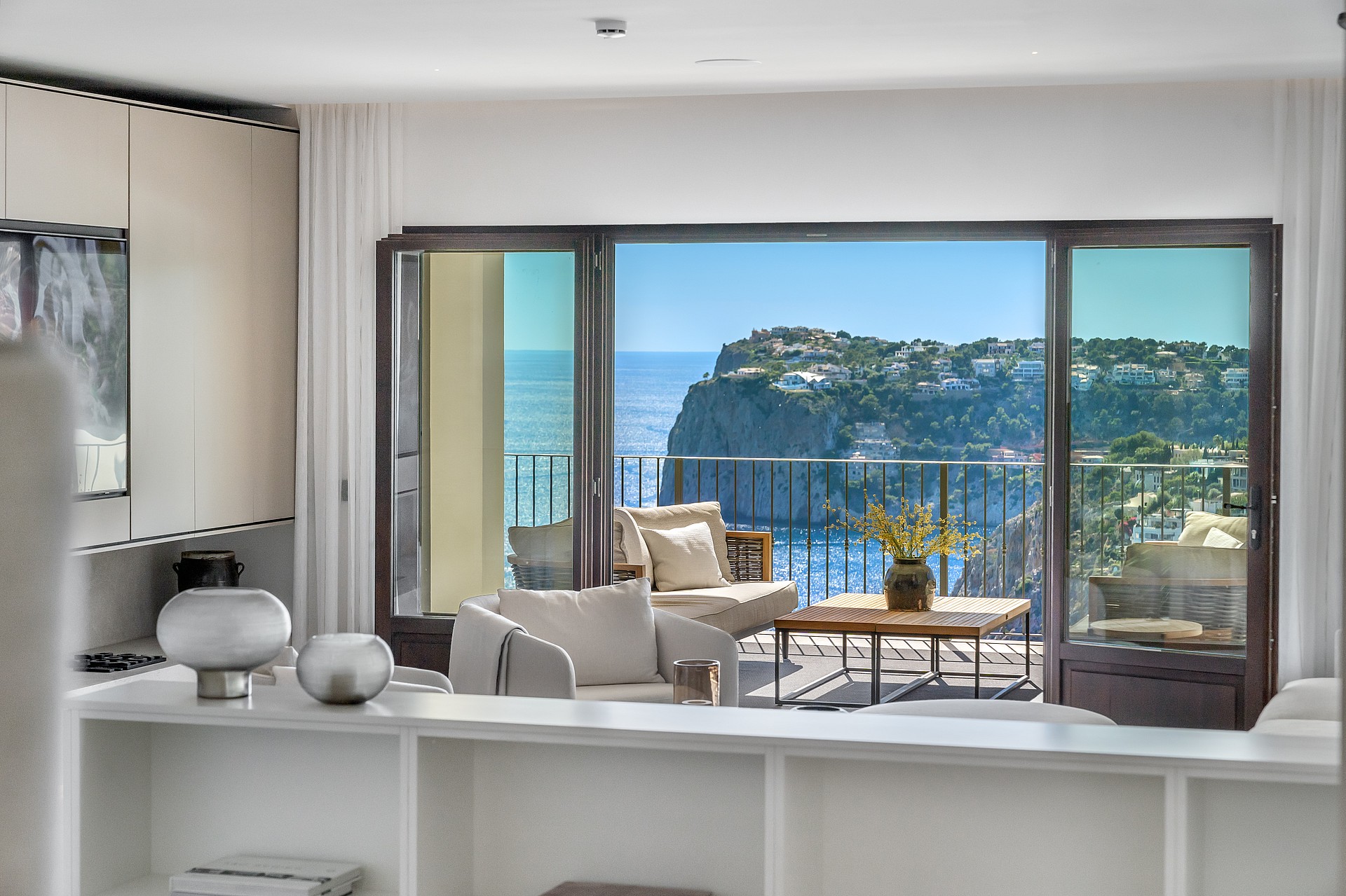 Interior view through dark-framed glass doors to a furnished terrace overlooking a coastal cliff and the blue sea.