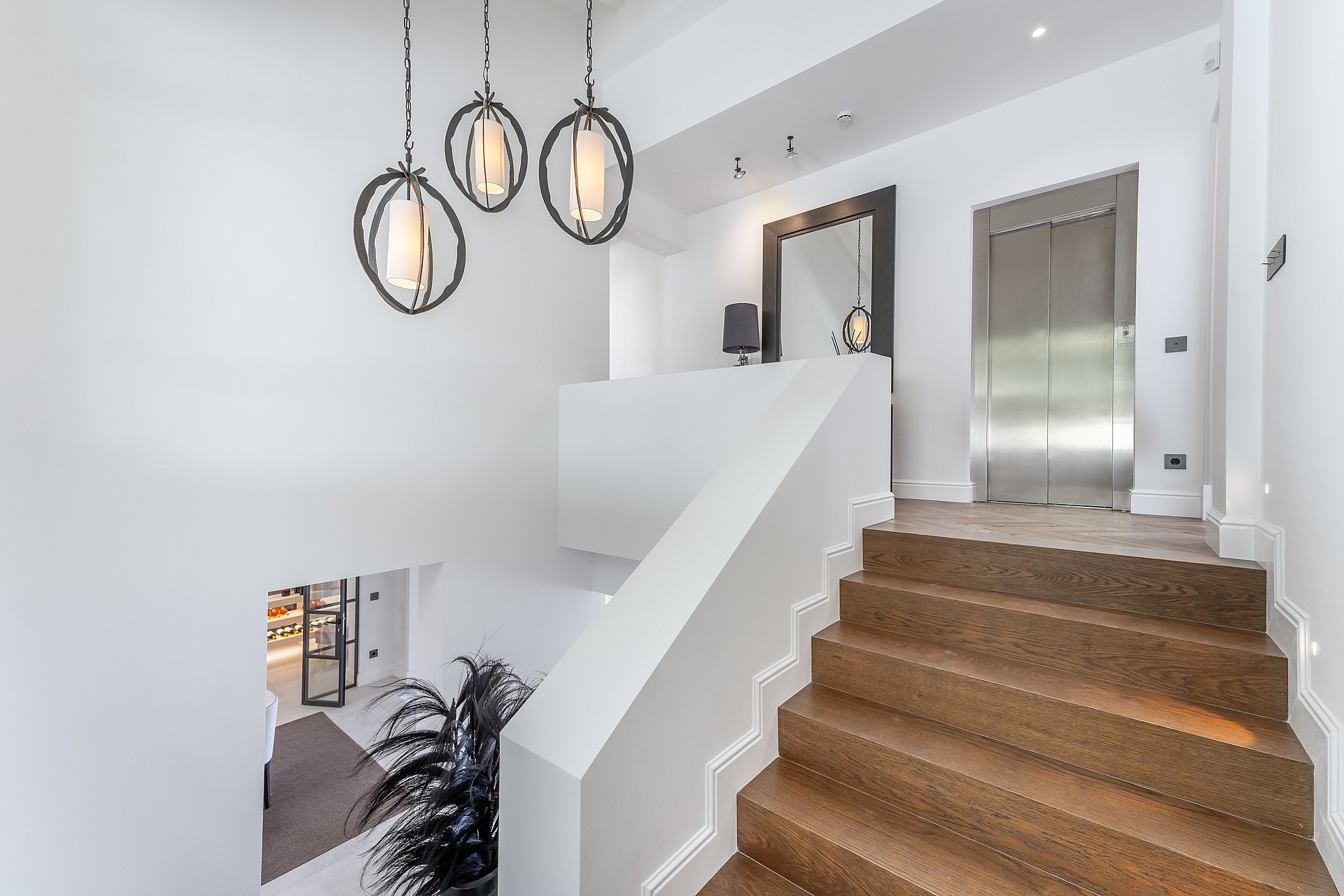 A white staircase with oak steps sits adjacent to a brushed steel elevator and three circular pendant lights.