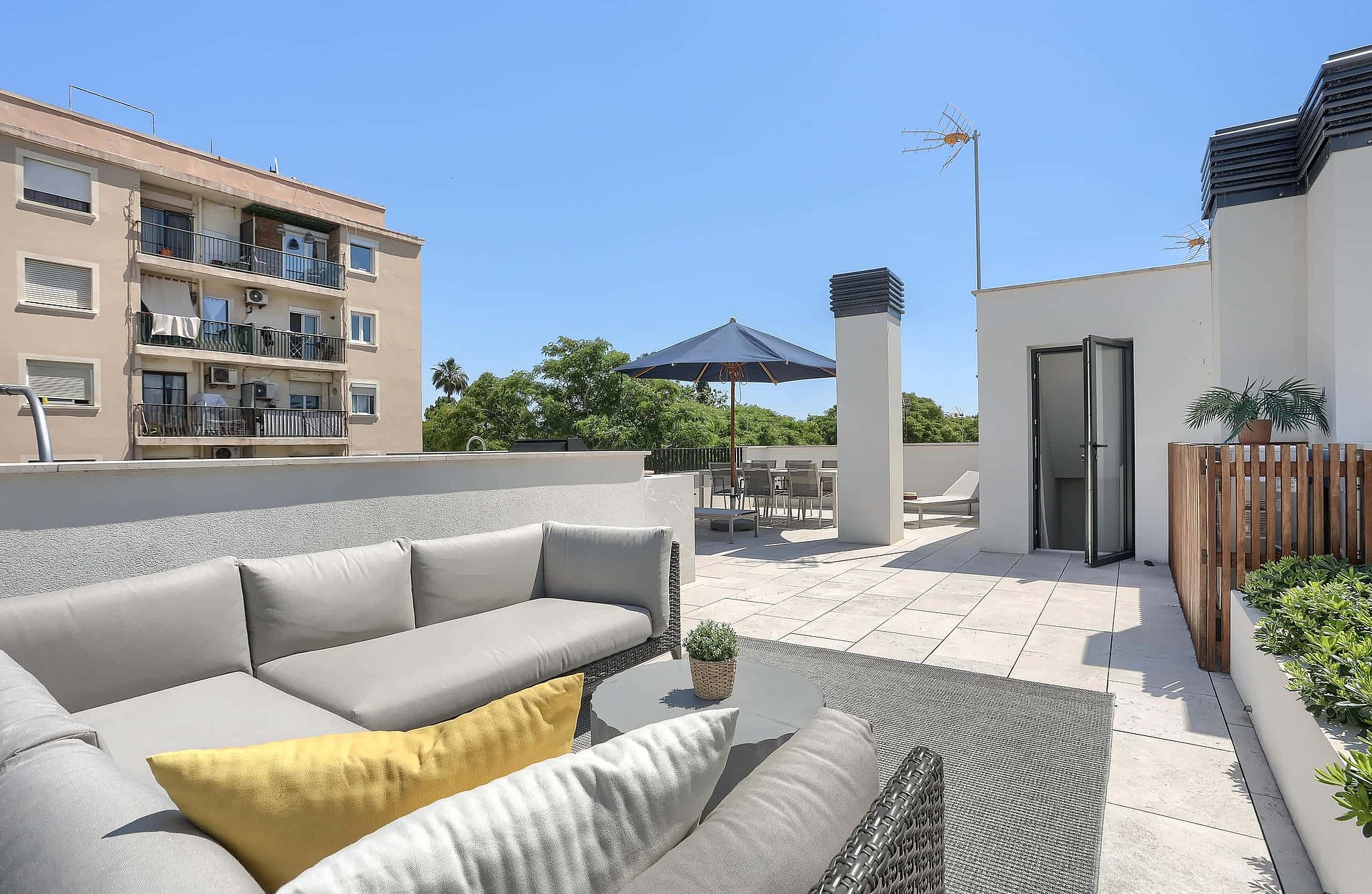 A spacious rooftop terrace featuring a light grey sectional sofa with yellow cushions, white stone flooring, and a dining area with a navy blue parasol under a clear blue sky.