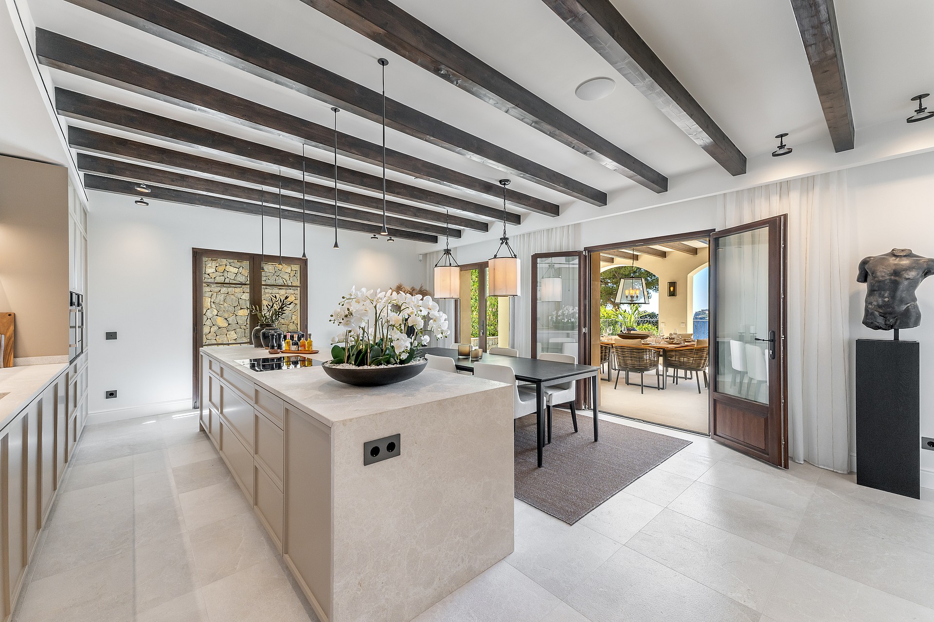 A wide stone kitchen island and dark ceiling beams in an open-plan room opening onto a sunlit outdoor dining terrace.