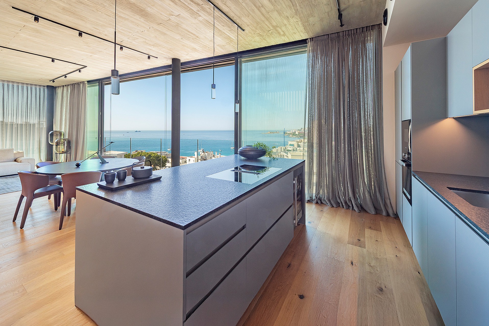 Modern kitchen island with dark stone countertop, light wood flooring, and floor-to-ceiling glass windows overlooking the sea.