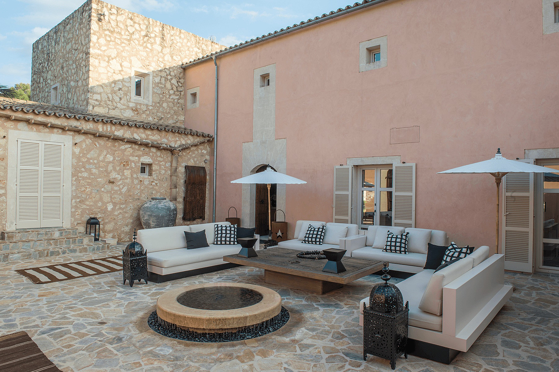 An outdoor stone courtyard of a Mallorcan finca featuring cream-colored sofas, a circular fire pit, and rustic stone walls.