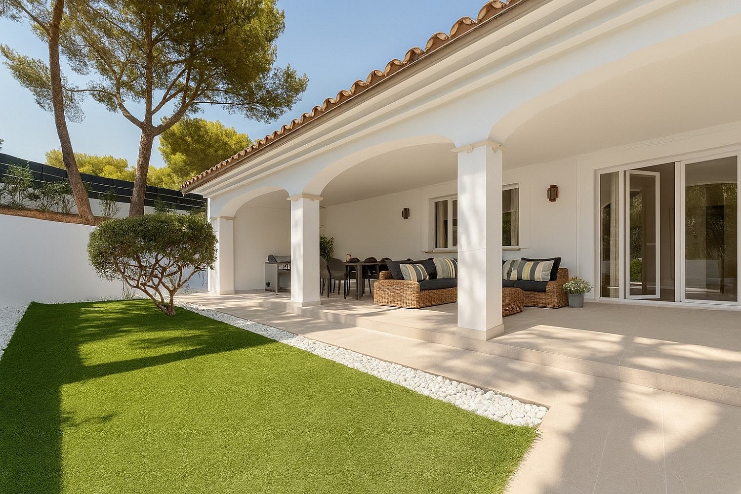 Covered outdoor terrace with white arched columns, dining table and wicker sofa opening onto lawn and mature pines