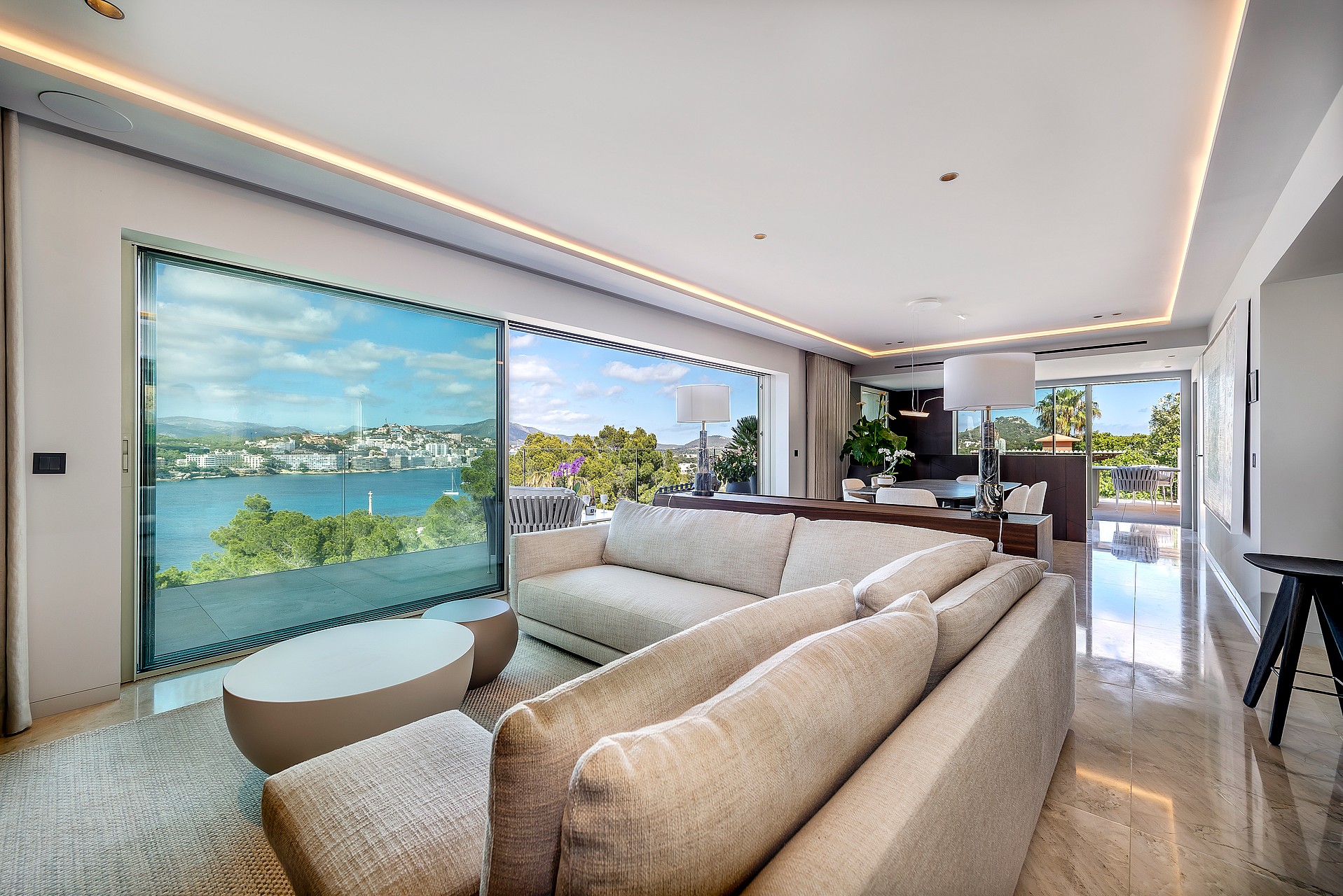 A contemporary open-plan living room featuring a large beige sectional sofa, polished stone floors, and floor-to-ceiling glass sliding doors opening to a lush garden view.