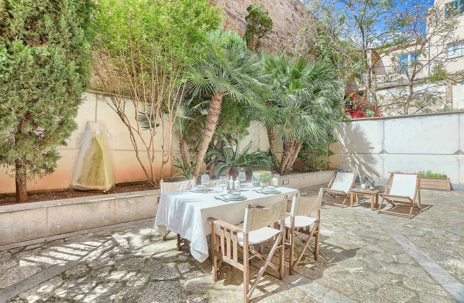 An outdoor dining table with white tablecloth and director chairs set on a gravel terrace shaded by mature trees.