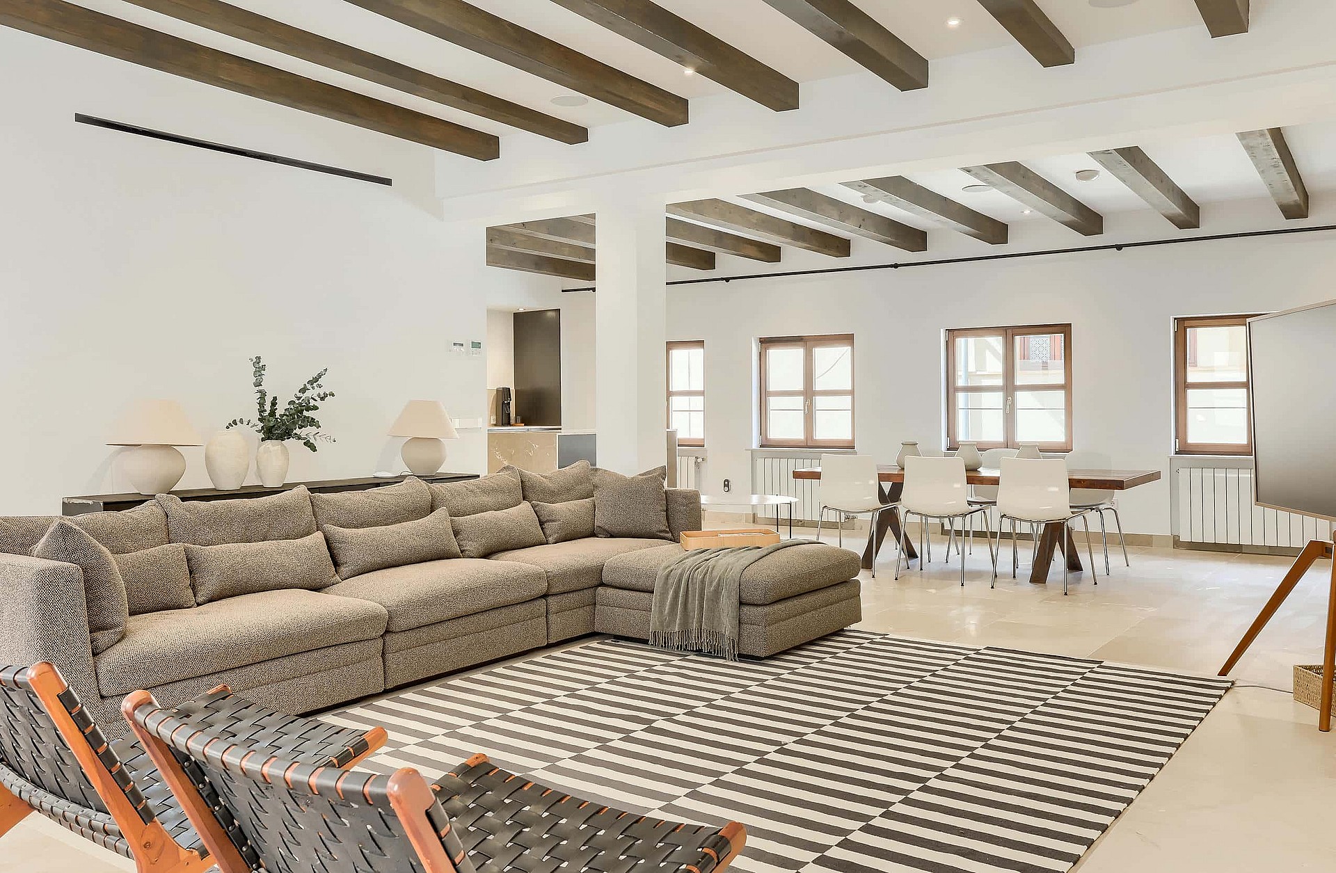 A spacious living room featuring a large patterned sofa, exposed dark wood ceiling beams, and a dining area in the background.