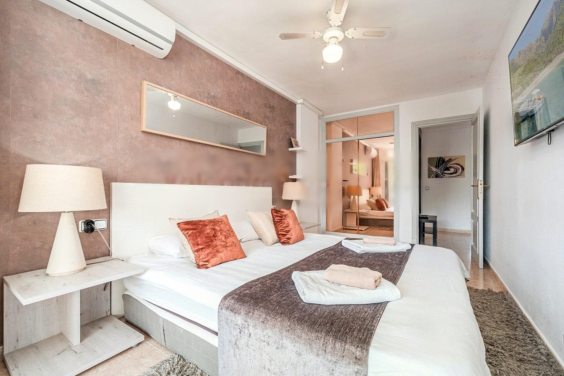 A bedroom featuring a textured dusty rose accent wall, a white bed with orange decorative pillows, a ceiling fan, and a view into a secondary room through an open doorway.