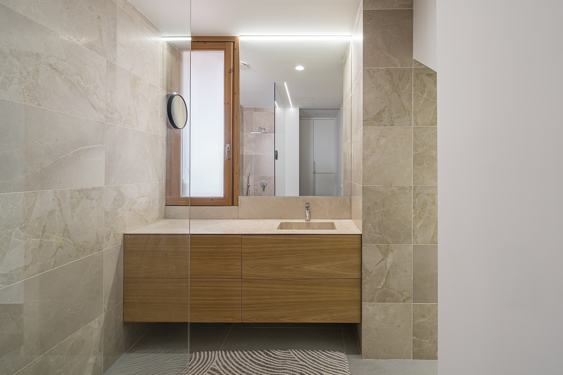 A modern bathroom featuring a wall-mounted wooden vanity, large rectangular mirror, and beige natural stone wall tiles.