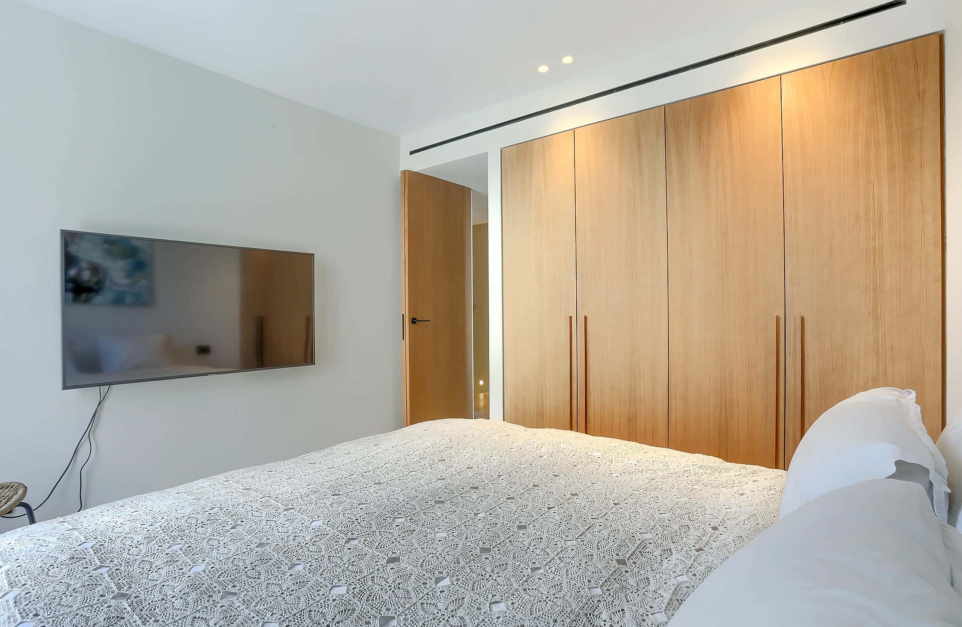 Contemporary bedroom featuring a large oak wardrobe wall, wall-mounted television, and neutral minimalist decor.