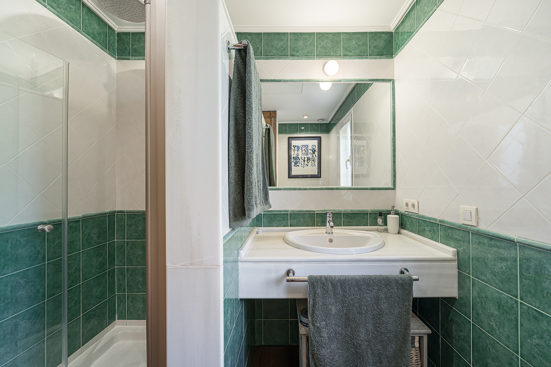 A bright bathroom with white and green square wall tiles, a glass-enclosed shower, a white vanity with a sink, and a rectangular mirror.