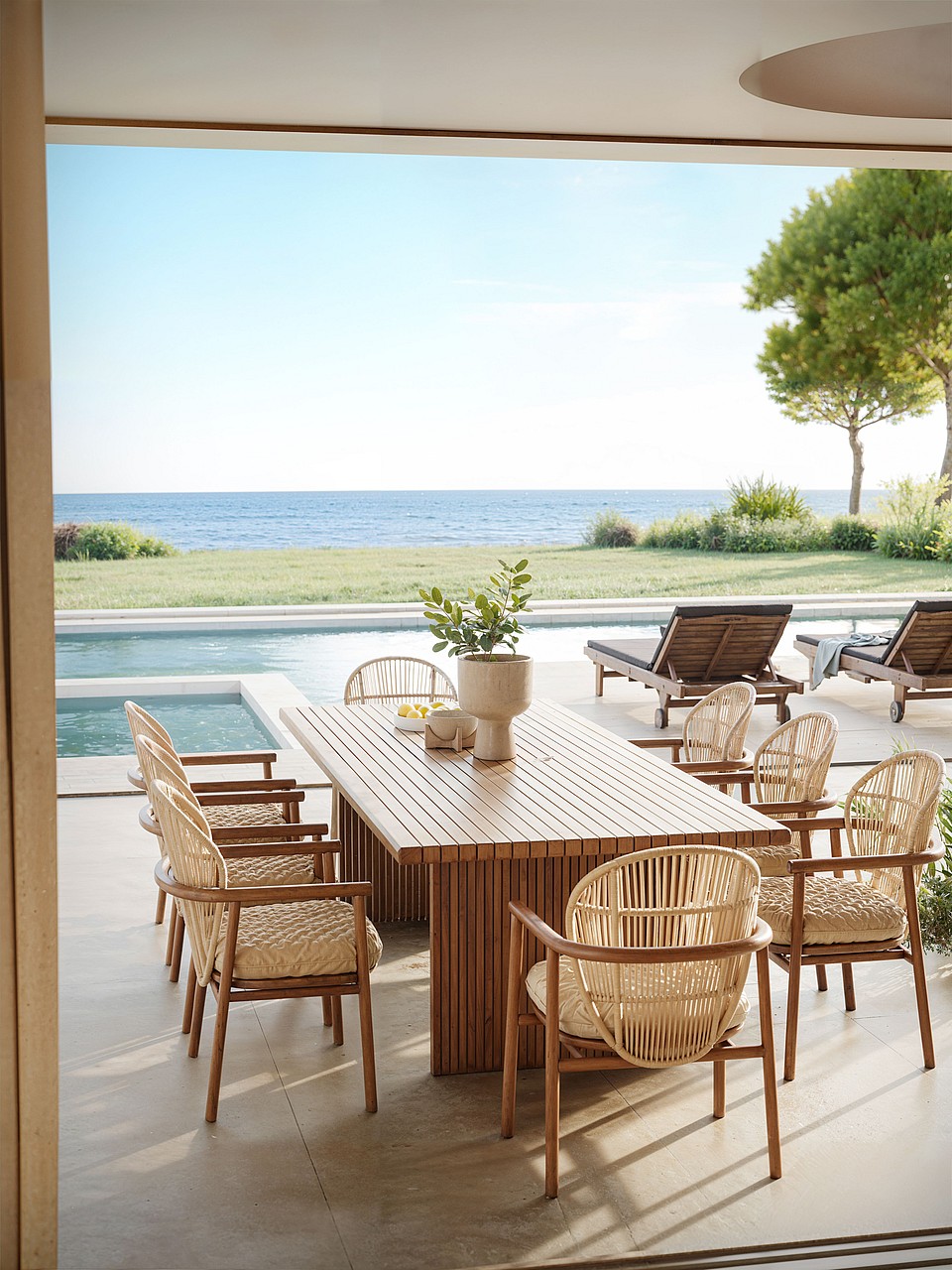 A slatted wood outdoor dining table with wicker chairs on a covered terrace, facing a swimming pool and the Mediterranean sea.