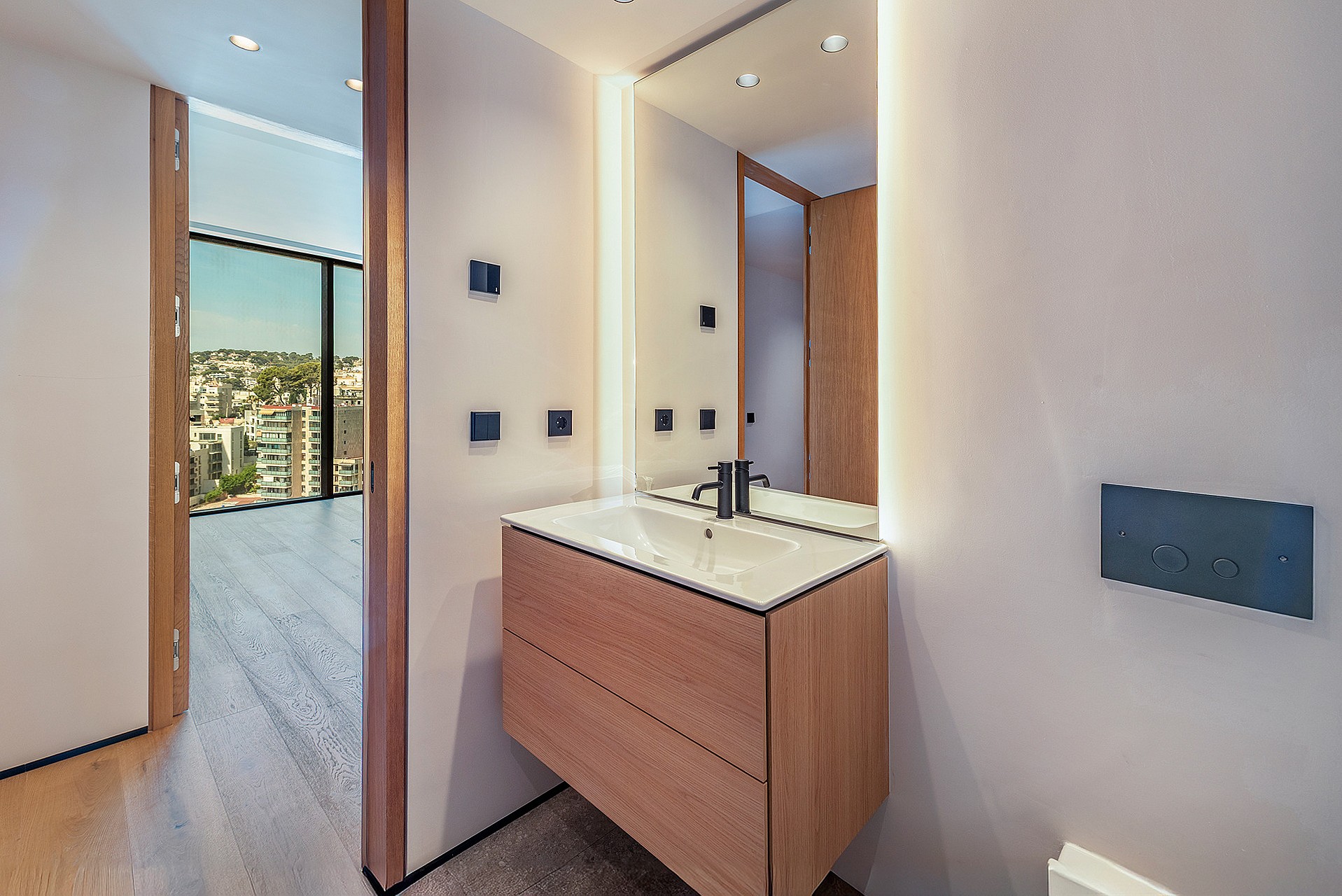 A bathroom vanity with a wooden cabinet and white countertop, featuring integrated mirror lighting and a view of the city.