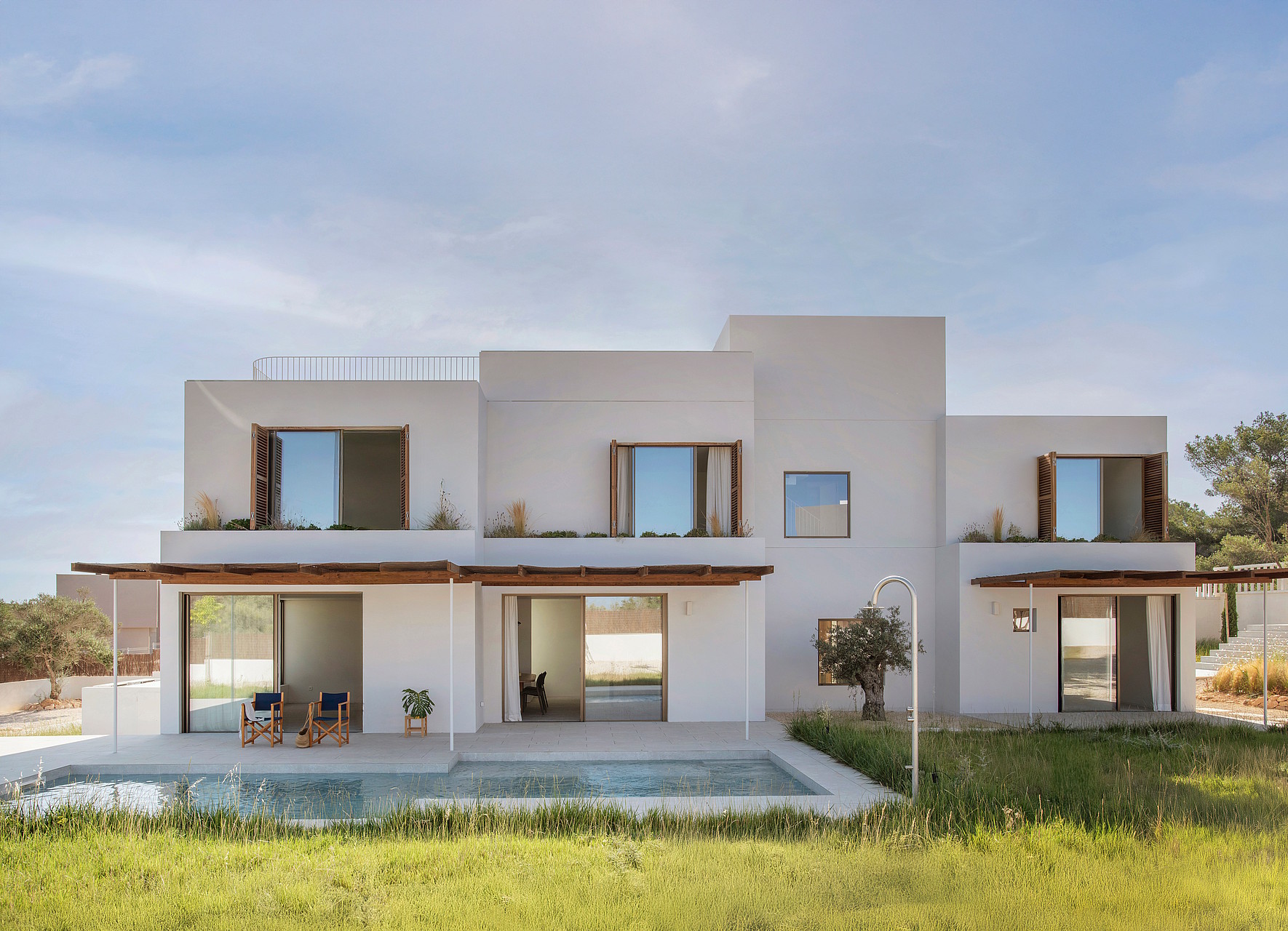 Modern white two-story villa with wooden shutters and a rectangular pool reflecting the sky, bordered by green grass.