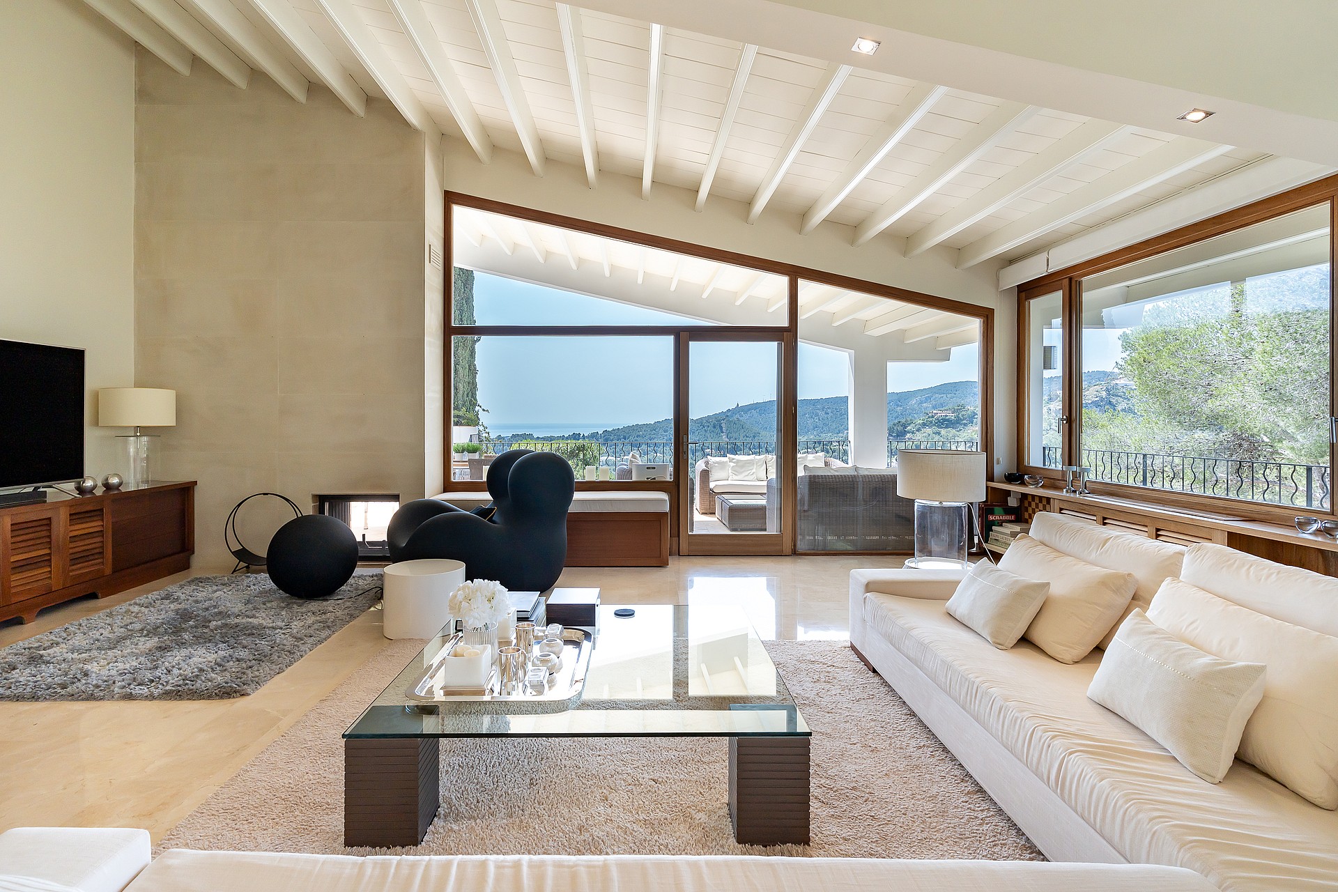 Spacious villa living room with white painted beams, fireplace, marble floors, designer seating and glazed wall with sea views