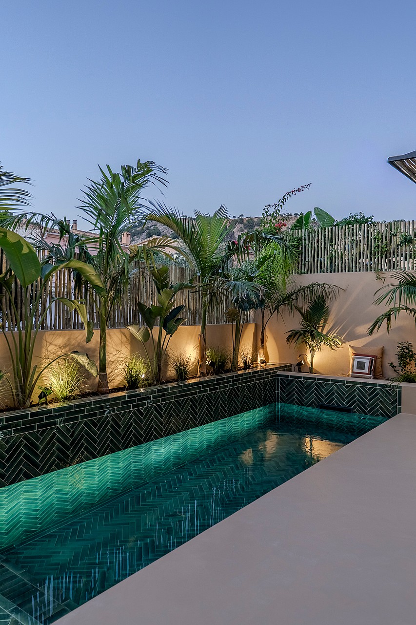 A rectangular outdoor swimming pool featuring dark green patterned tiles, integrated lighting, and tropical landscaping.