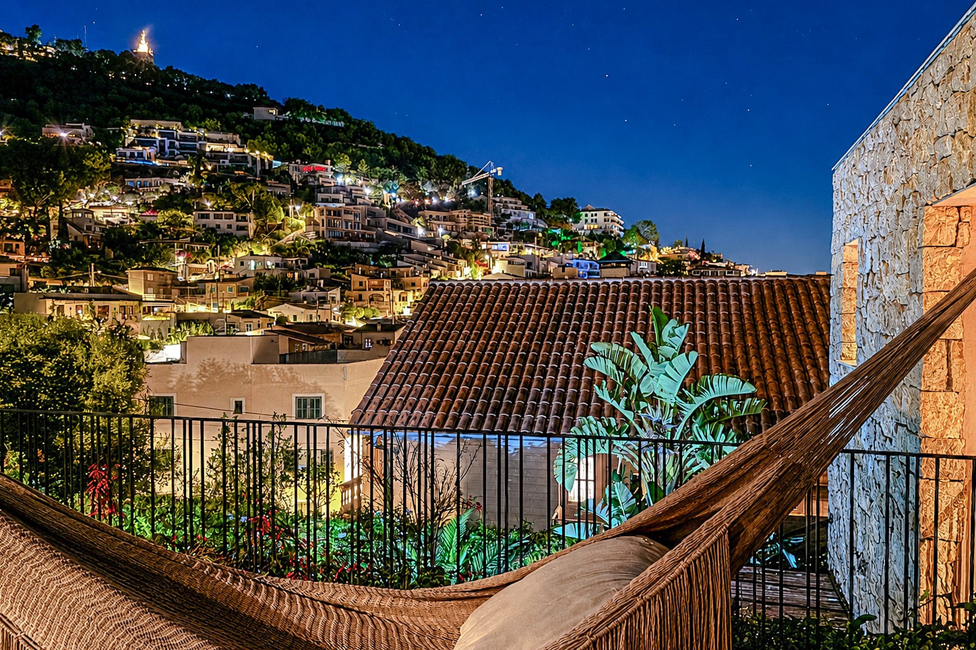 A stone-walled terrace at twilight, featuring a hammock, metal railing, and a view of hillside town lights in Genova.