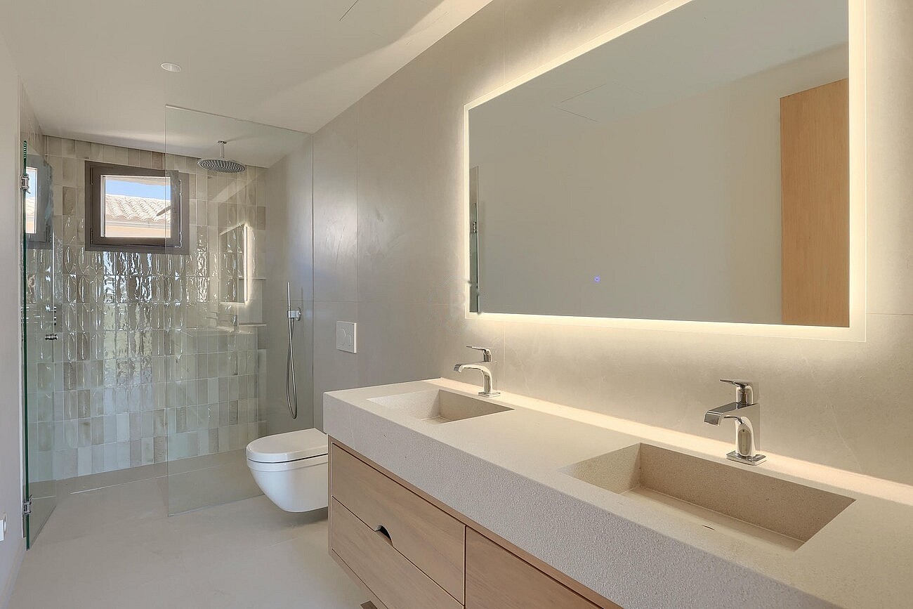 Contemporary bathroom with a light-toned wooden vanity, integrated stone double sink, large backlit mirror, and glass-enclosed shower.
