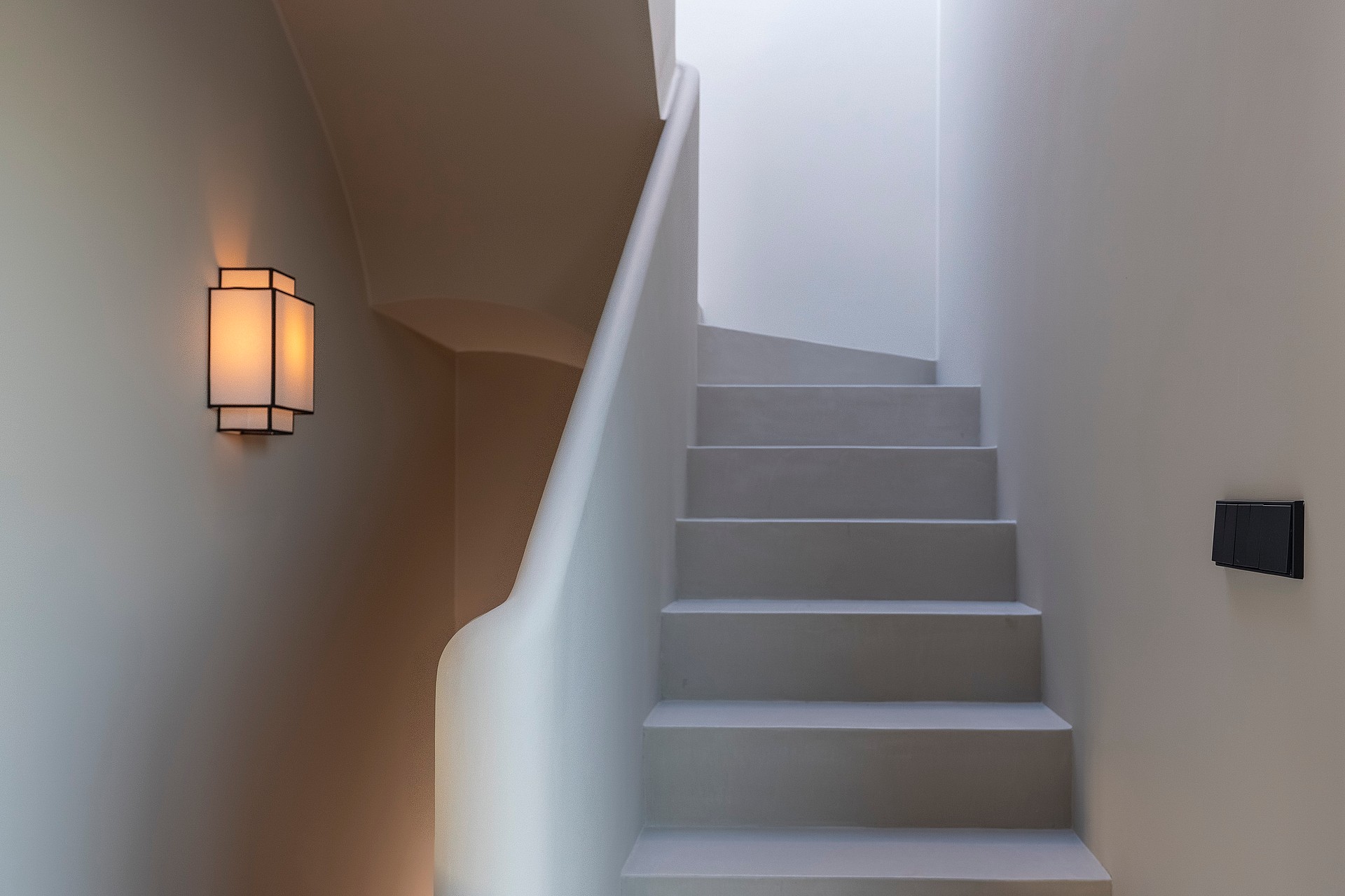 A minimalist microcement staircase with a curved balustrade, illuminated by a warm wall sconce and soft natural light.