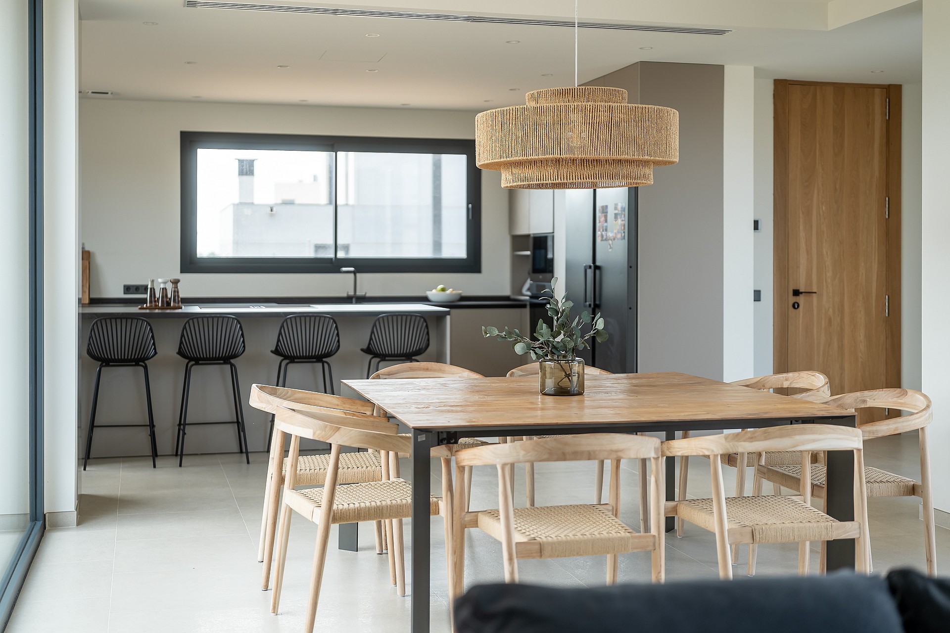 A dining space featuring a large square wooden table with woven chairs, a textured light fixture, and a modern kitchen island with black bar stools in the background.