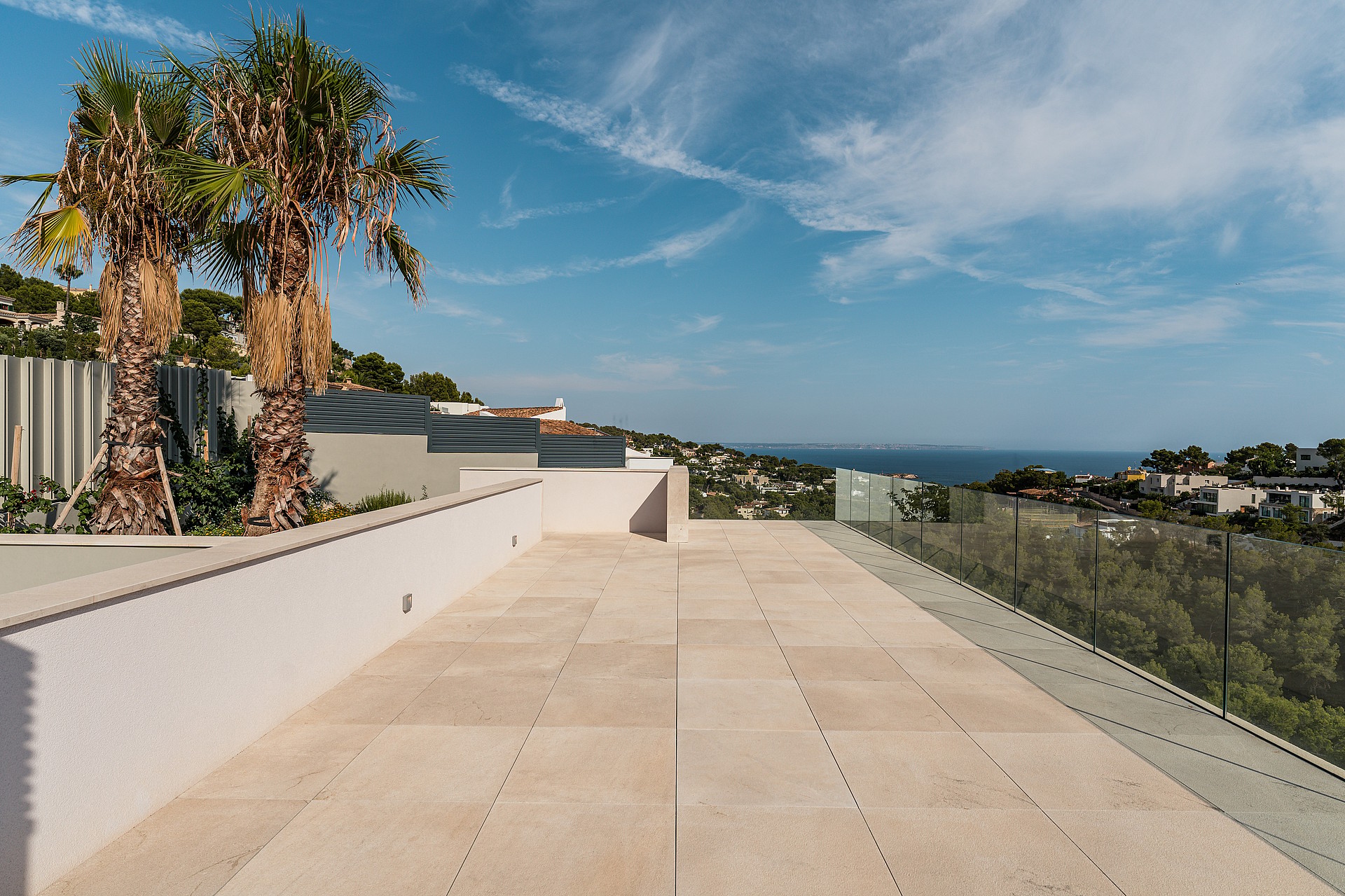 A wide outdoor stone terrace with palm trees, white perimeter walls, and glass railings overlooking a distant coastal landscape.