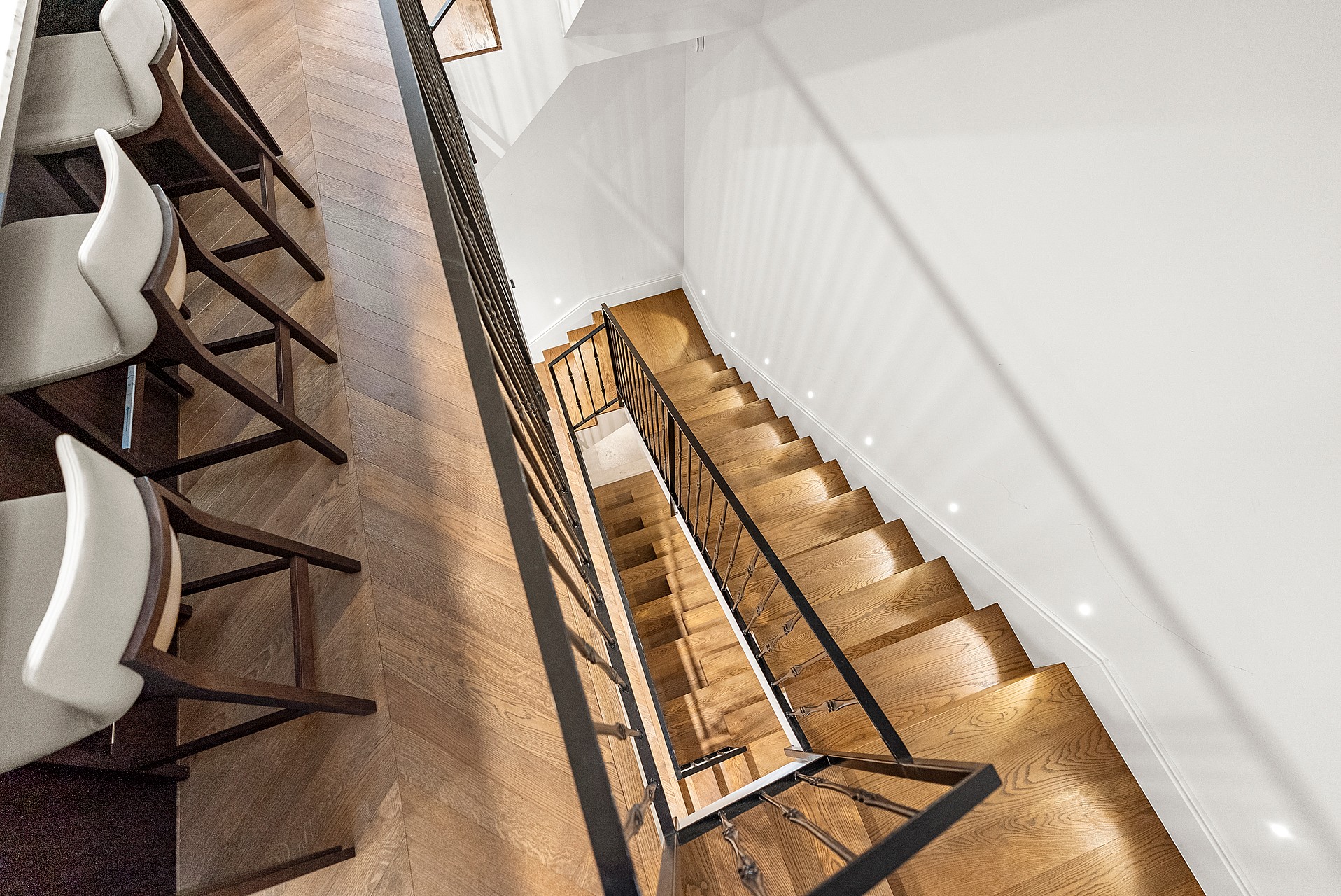 High-angle view of chevron wood flooring, leather-seated bar stools, and a black metal staircase railing.