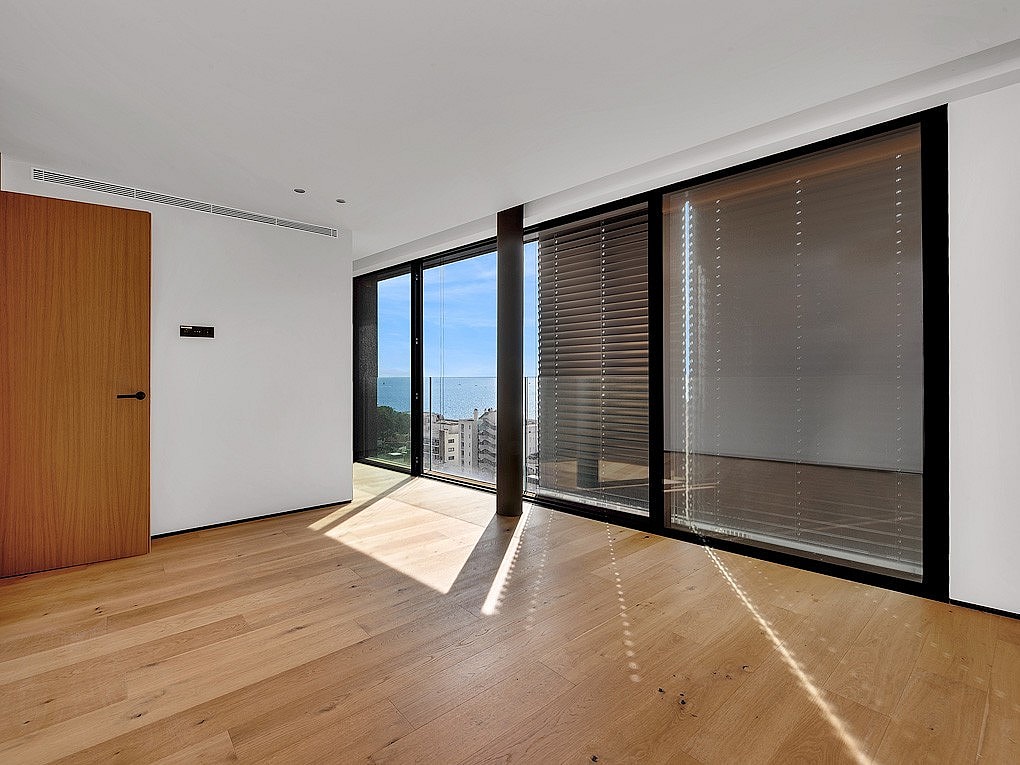 A minimalist bedroom featuring light oak flooring, white walls, and large floor-to-ceiling sliding glass doors that open to a balcony with sea views.