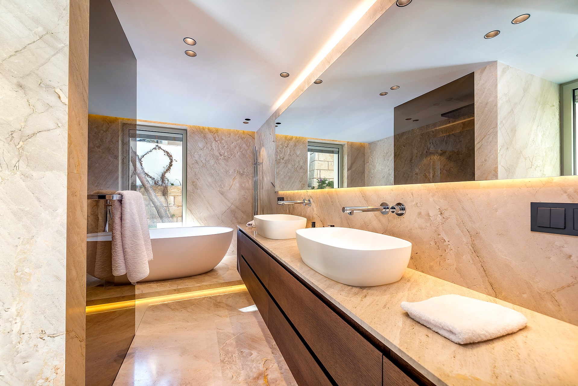 A modern bathroom with dual oval vessel sinks on a wooden vanity, a freestanding white bathtub, and floor-to-ceiling beige travertine stone walls.