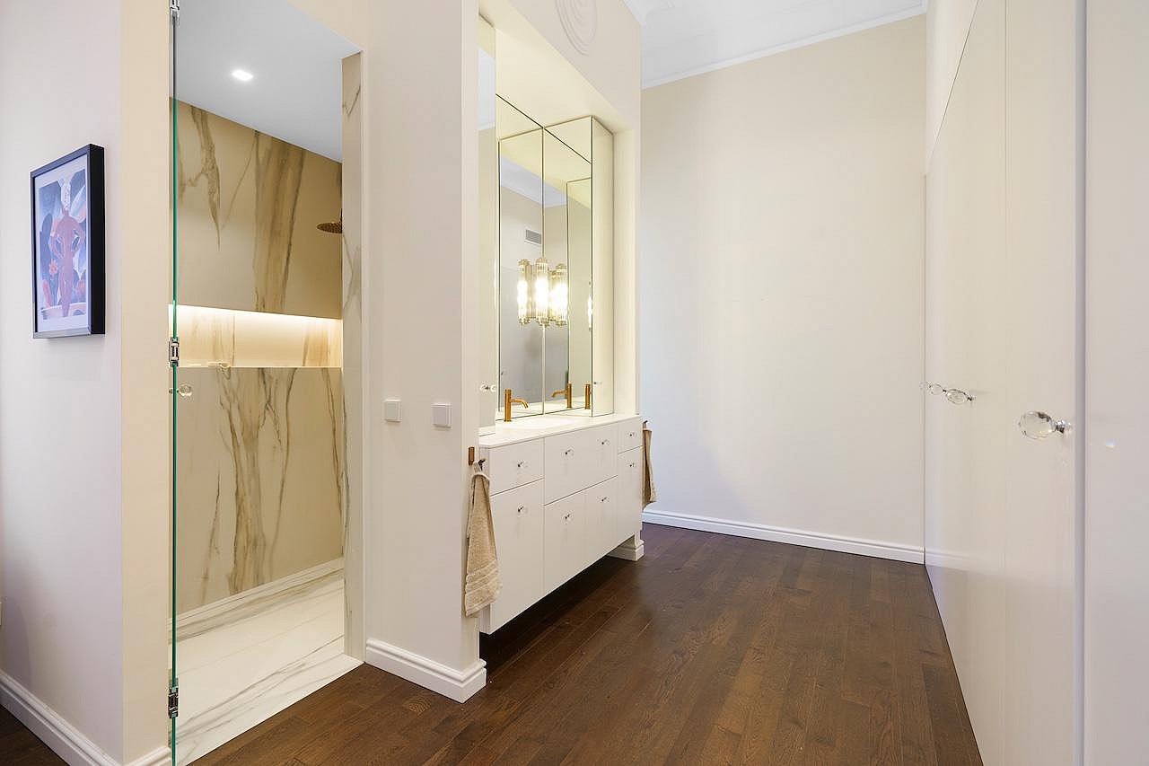 A modern bathroom featuring a walk-in shower with marble-veined walls, a white vanity with gold hardware, and dark wood flooring.