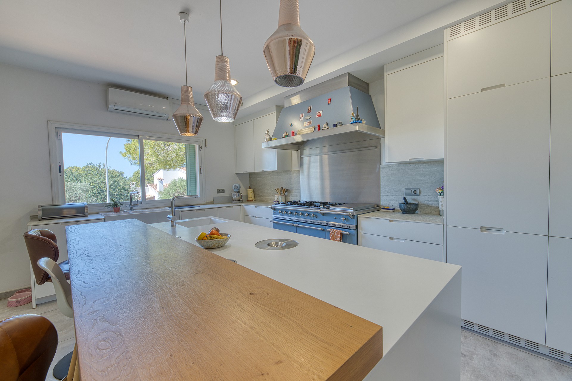 A modern white kitchen featuring a large central island with a wooden breakfast bar, stainless steel stove, and three glass pendant lights hanging above.