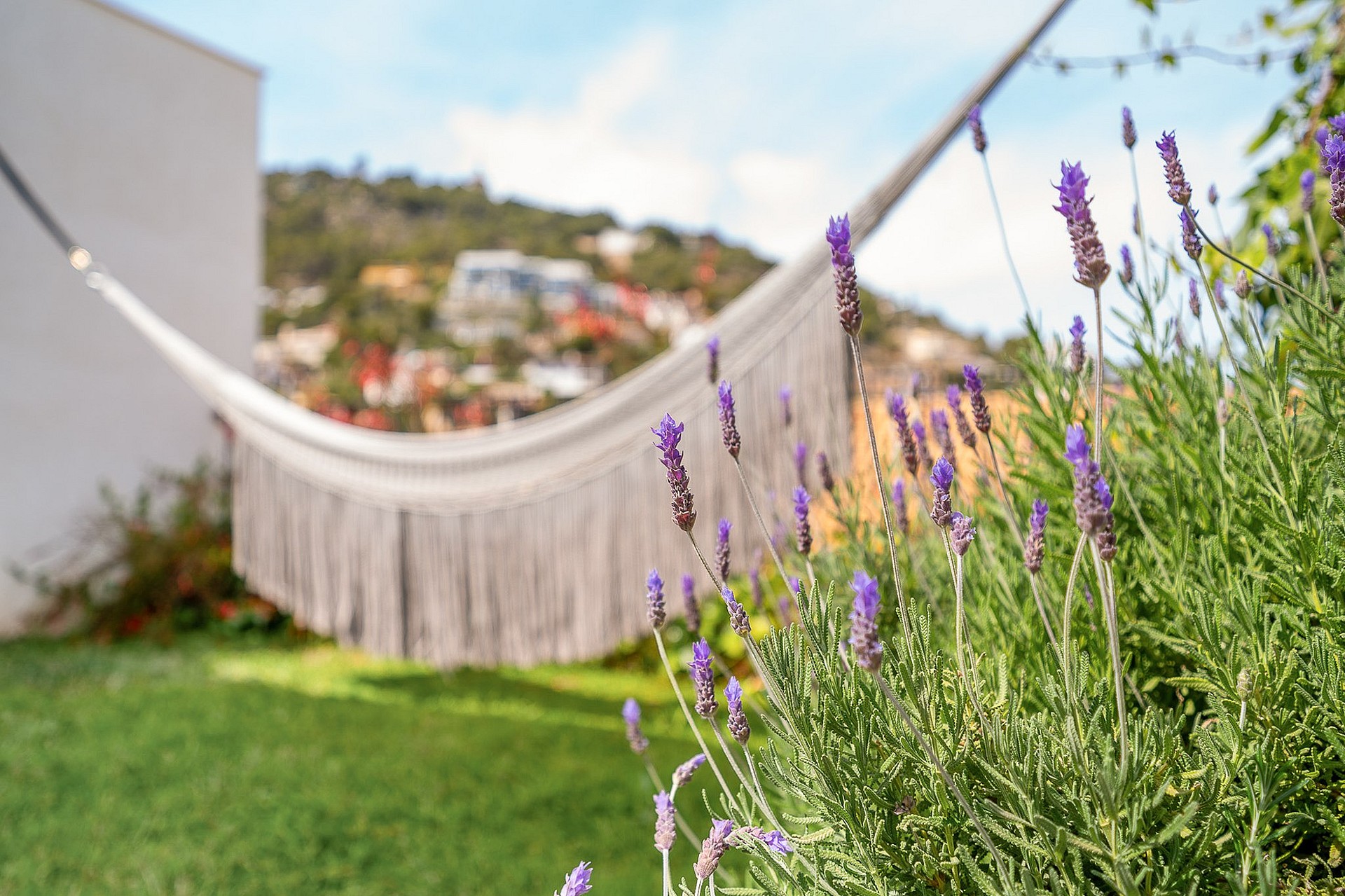 A white fringe hammock suspended in a garden, with blooming lavender in the foreground and a mountain hillside in the background.